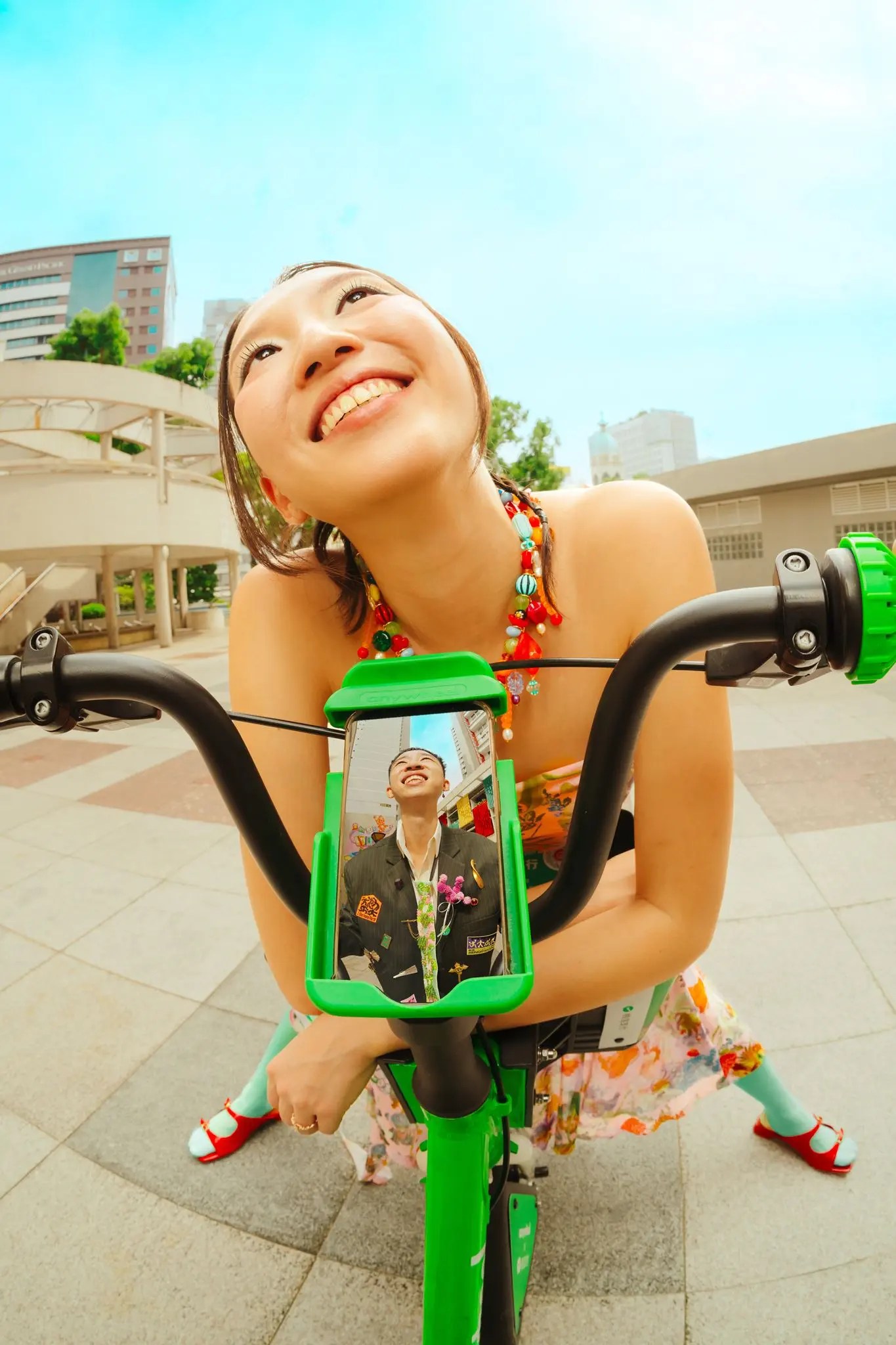 Rachel leaning over green bicycle handlebars laughing skyward in a chunky beaded necklace, a phone in the mount showing a photo of Rio