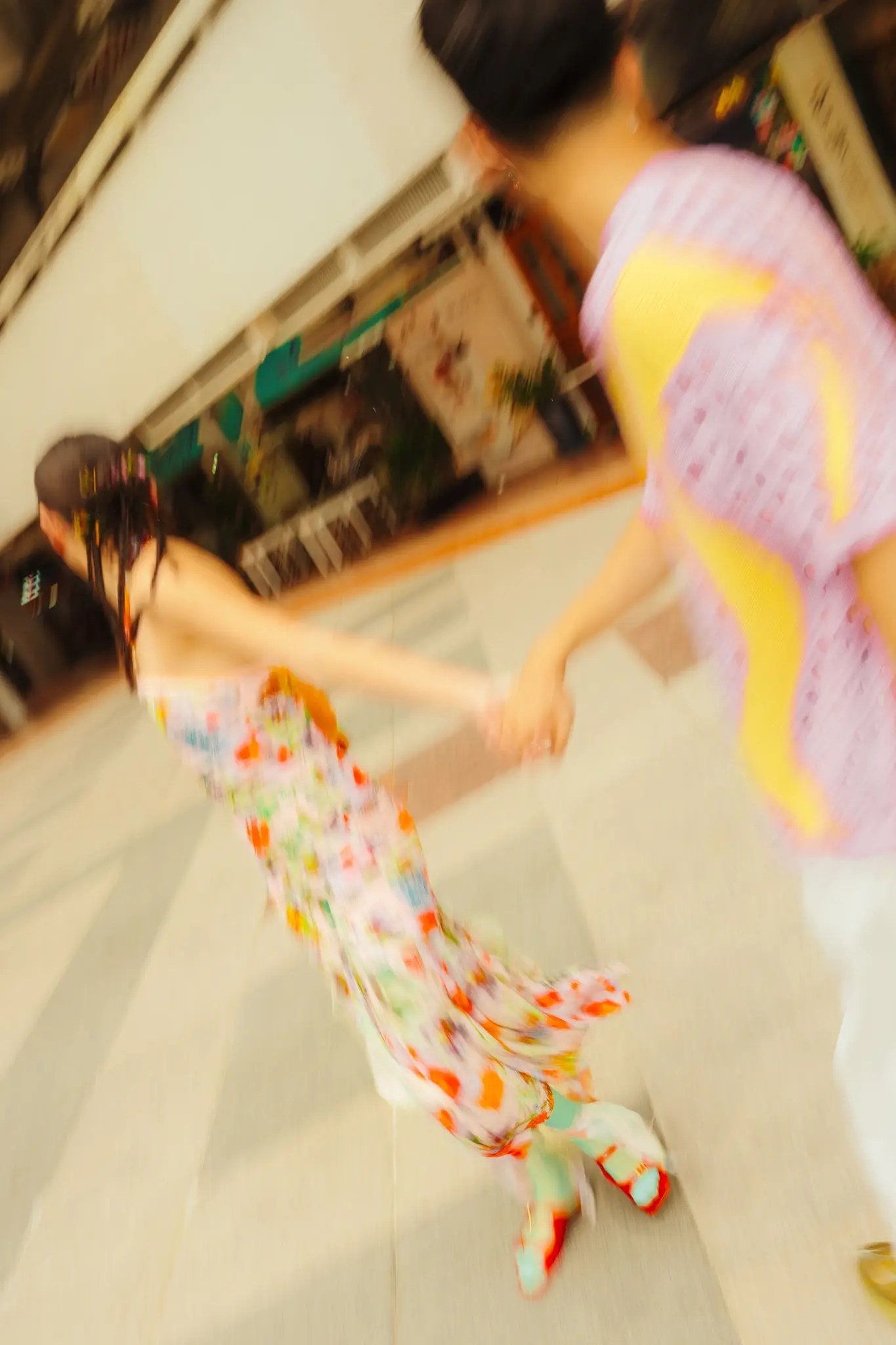 Motion-blurred shot of Rio and Rachel running hand in hand down a covered walkway, Rachel in a floral print dress and teal and red platform sandals