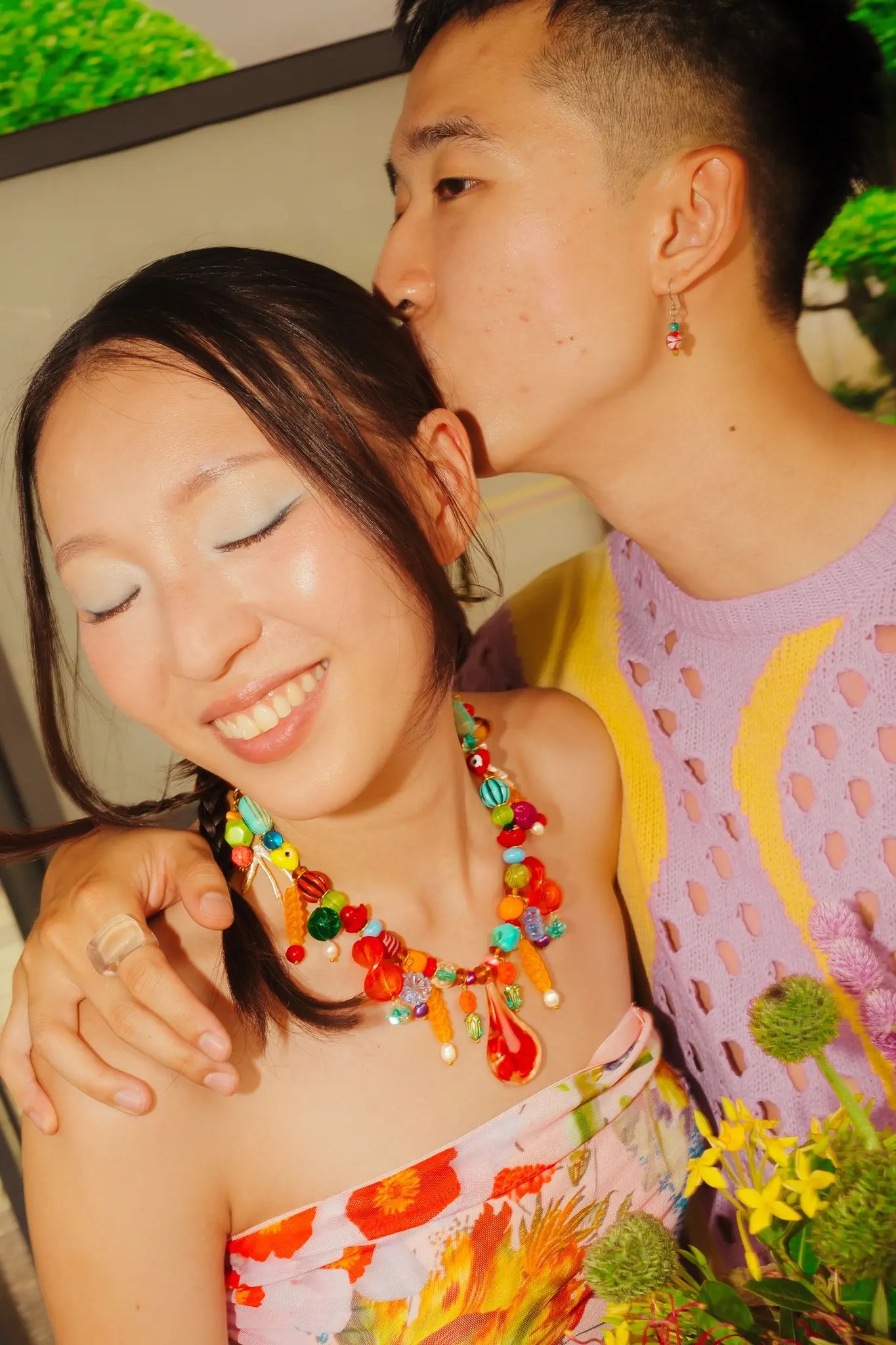 Rachel in a strapless floral Desigual dress and chunky multicolor beaded Inez Designs necklace smiling as Rio kisses Rachel's temple, yellow and green bouquet in hand