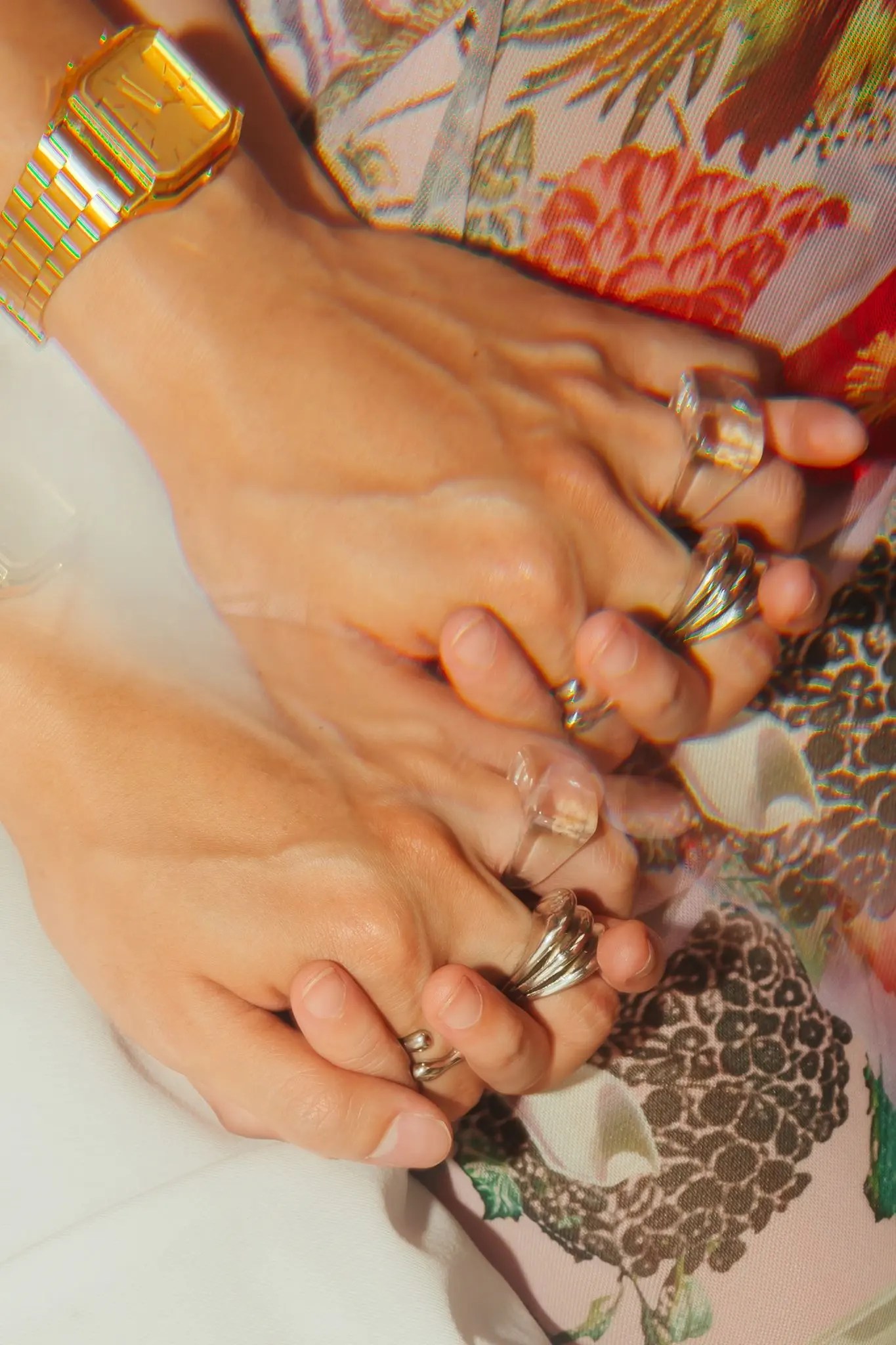 Close-up of Rio and Rachel's interlaced hands stacked with chunky silver sculptural rings and a gold digital watch against ivory fabric