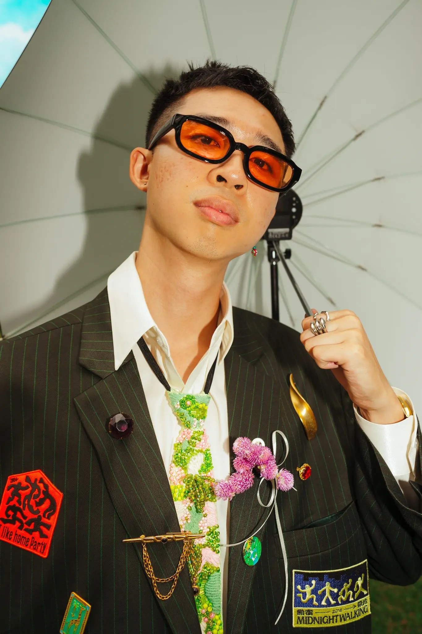 Rio in orange-lens sunglasses and a pinstripe jacket covered in embroidered patches, brooches by Nathanael Ng, and a beaded floral tie, posed against a white photo umbrella