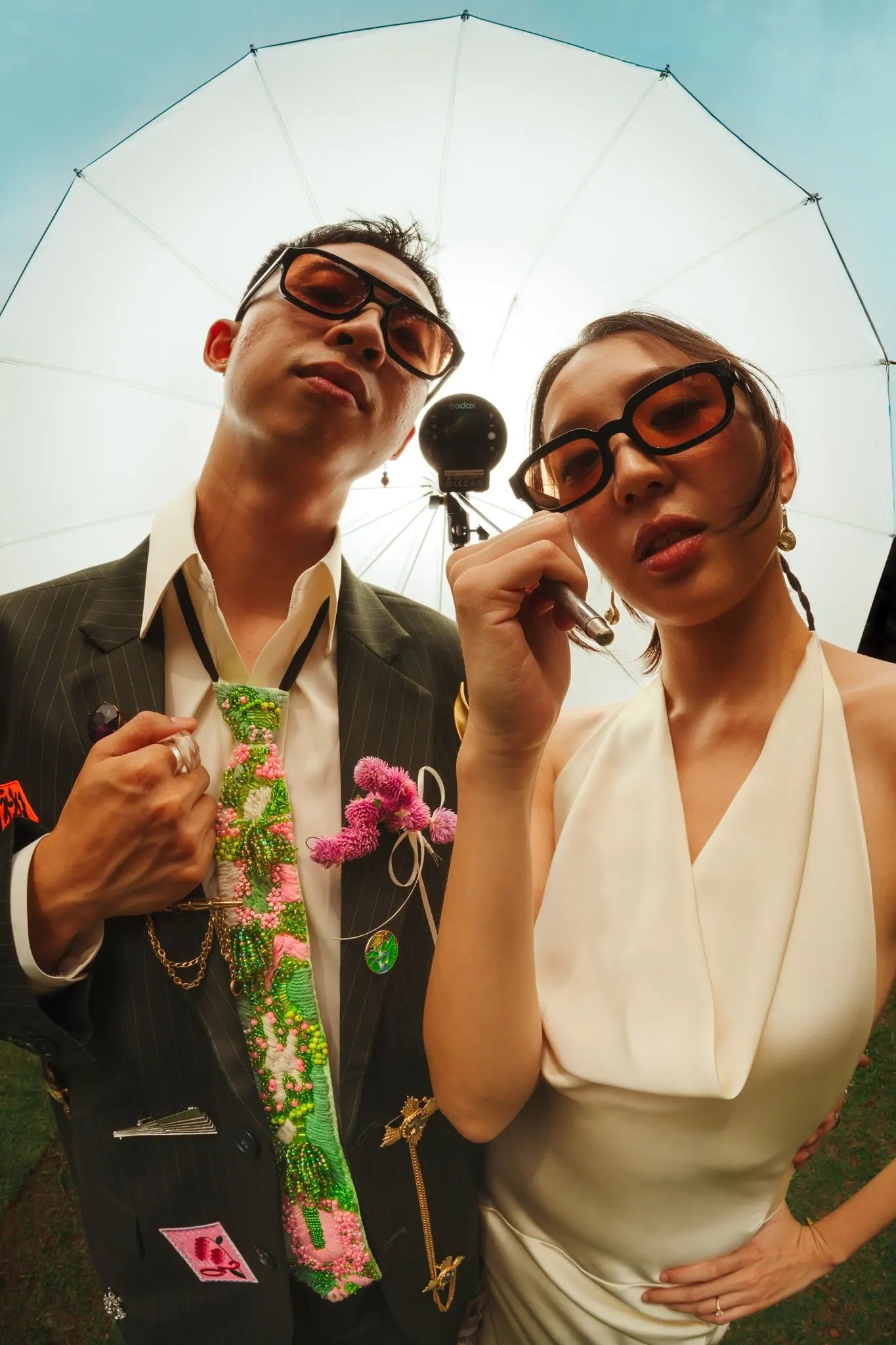 Low fish-eye of Rio and Rachel lowering their sunglasses toward the camera under a white photography umbrella and bright sky