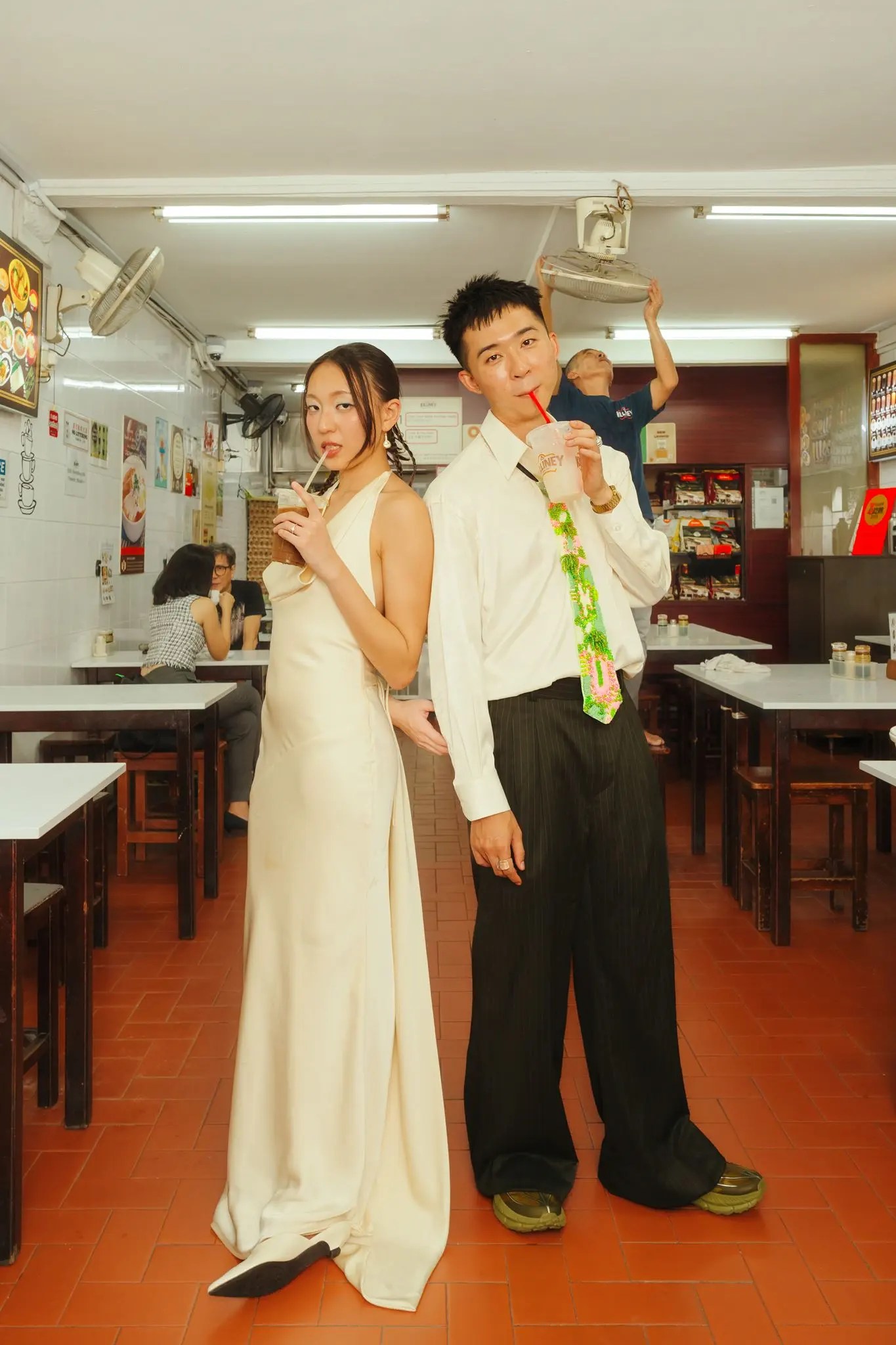 Rio and Rachel back to back sipping iced drinks through red straws in a kopitiam while a worker adjusts the ceiling fan behind them