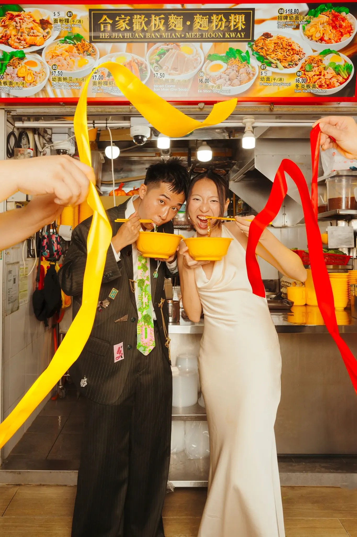 Rio and Rachel eating mee hoon kway at a Singapore hawker stall in an ivory satin halter gown and a pin-covered pinstripe suit, red and yellow ribbons in the air