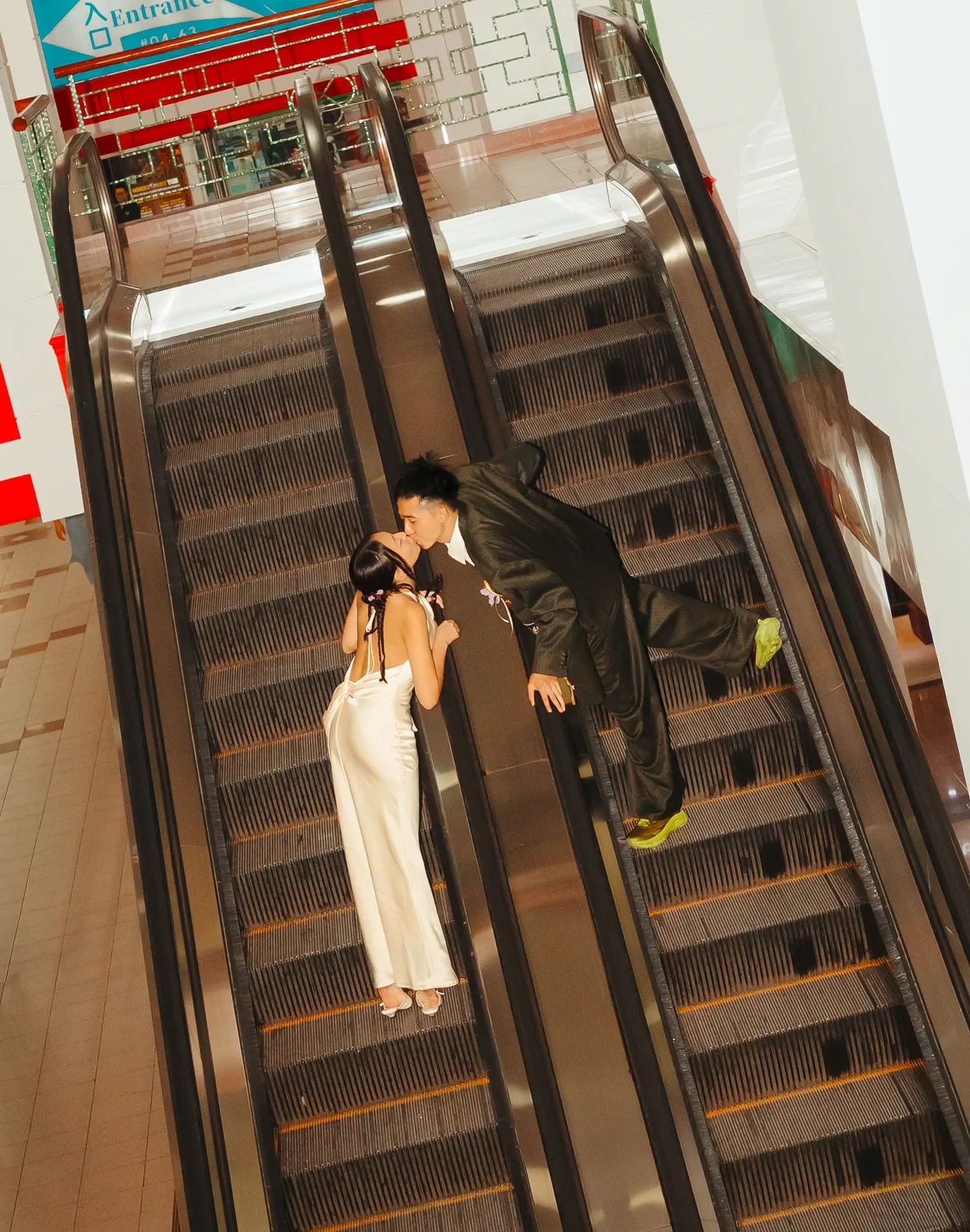 Overhead view of Rio leaning across a mall escalator divider to kiss Rachel, metal escalator treads filling the frame
