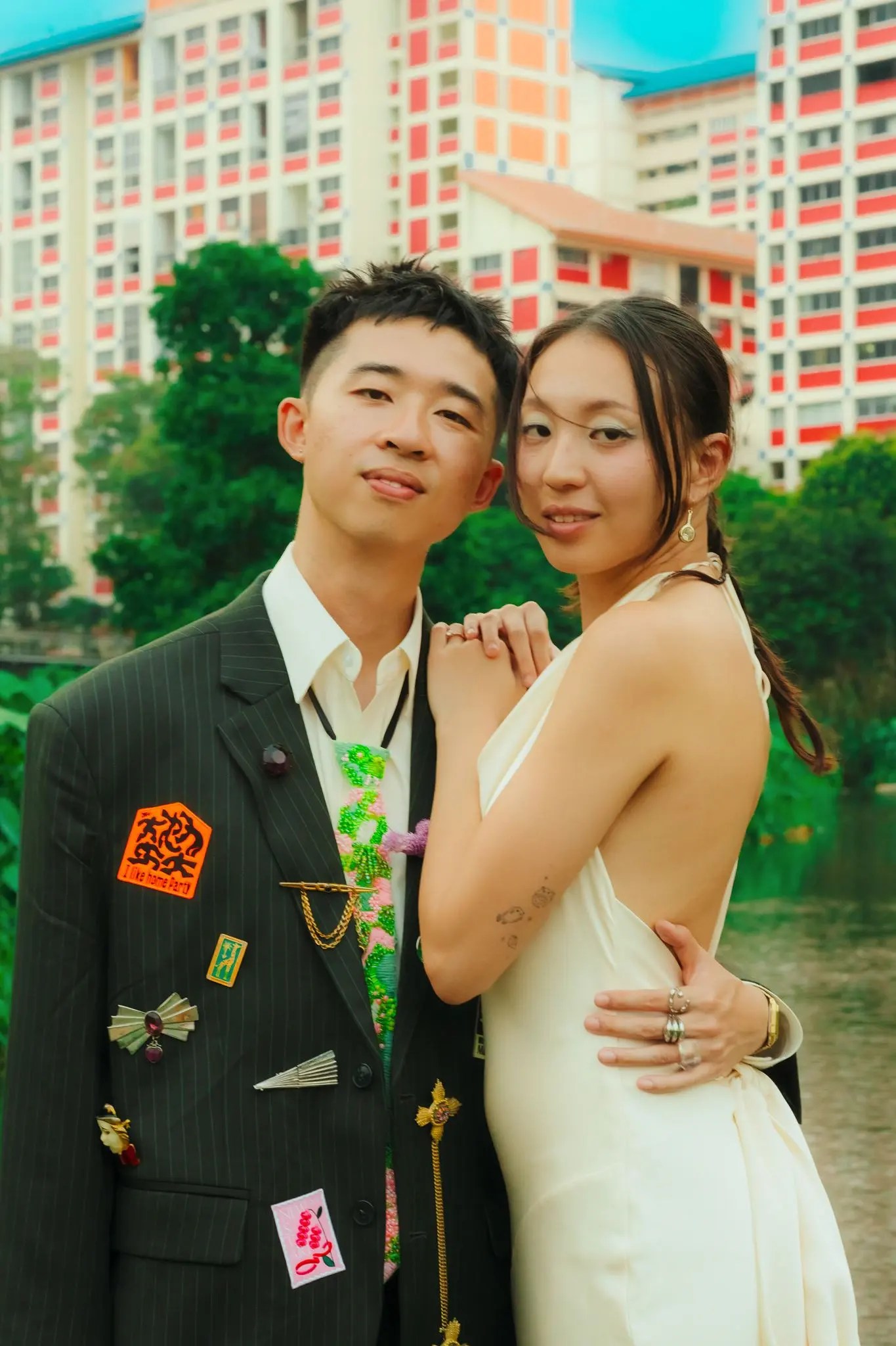 Couple embracing in front of pastel orange and cream HDB blocks and tropical greenery in Singapore, Rachel's open-back ivory gown turned to the camera