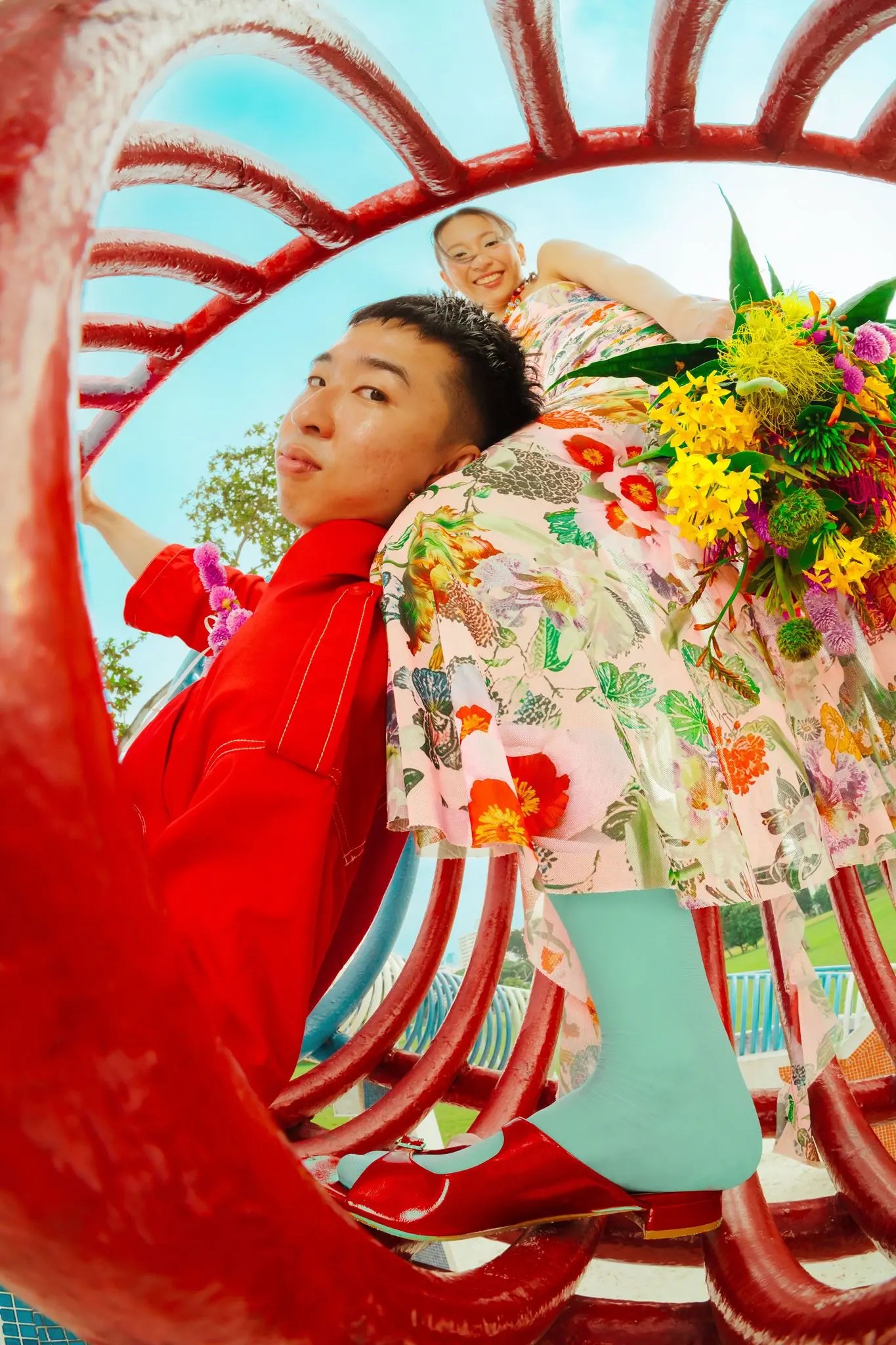 Rio in a red belted boilersuit framed by red tubular sculpture bars, Rachel draped above in a pink floral dress holding a Meteor Flowers bouquet of yellow blooms and purple globe amaranth