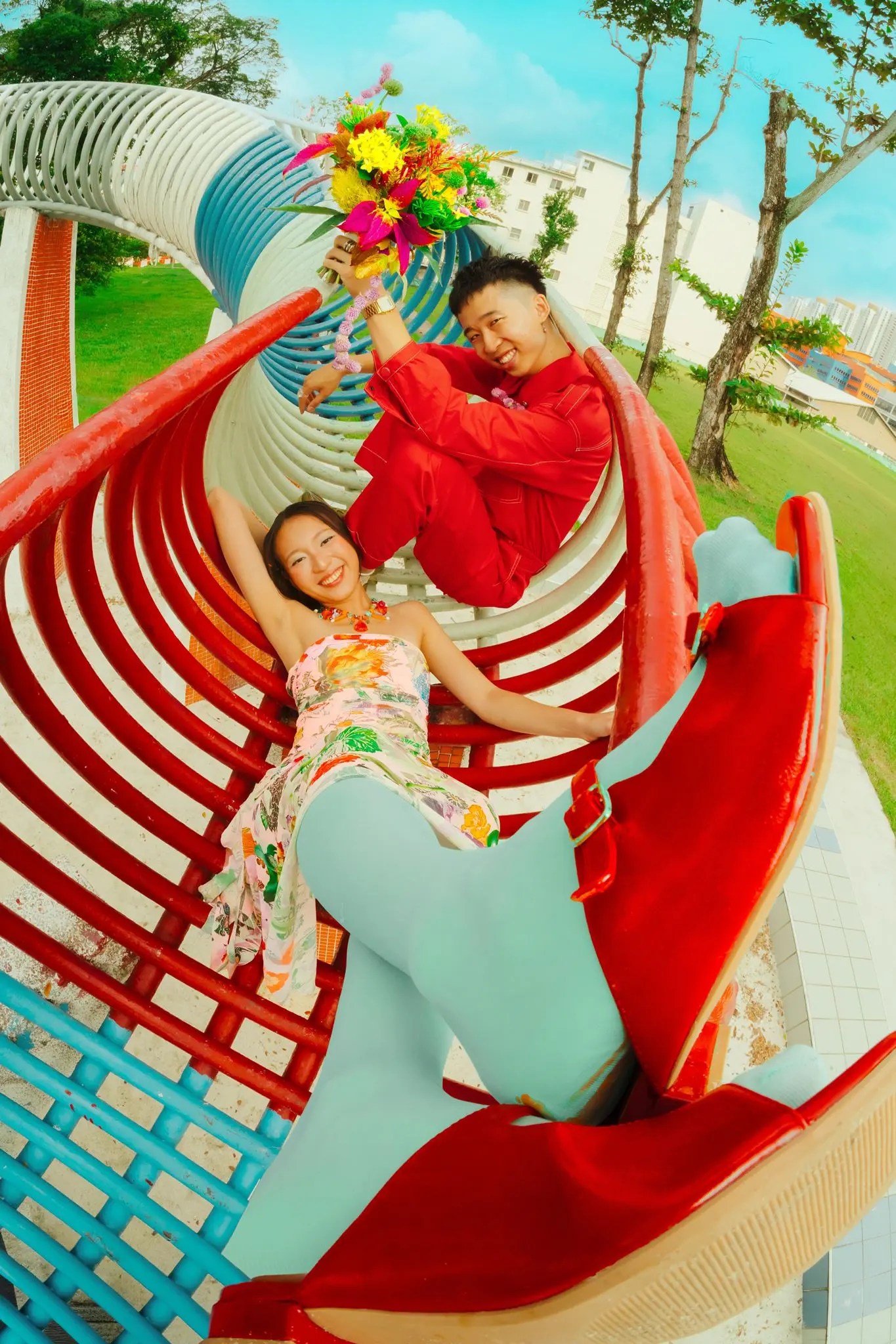 Wide fish-eye of Rachel reclining and Rio holding a wildflower bouquet aloft inside a red and pale blue curved sculpture, lawn and residential blocks behind