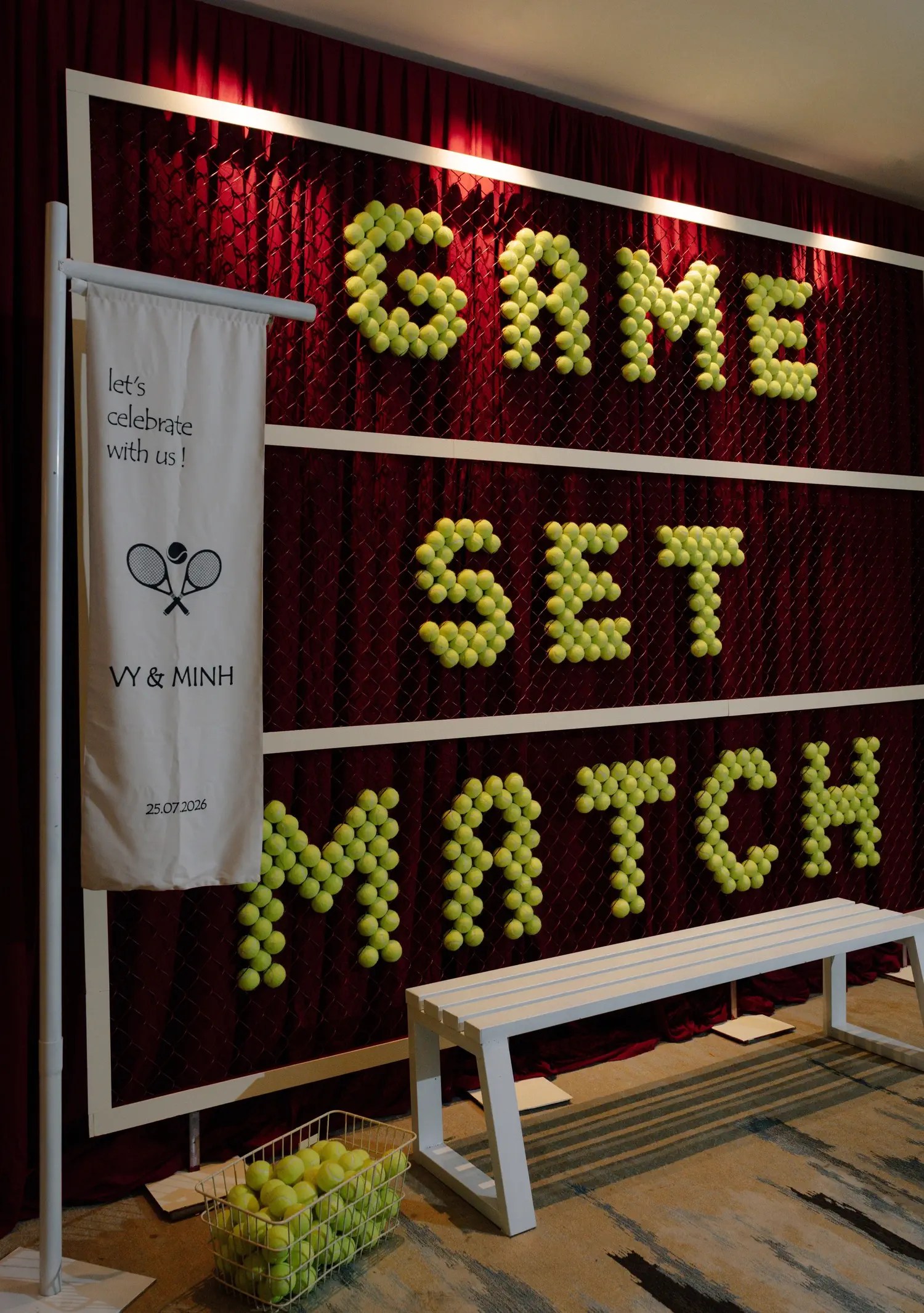Tennis-themed wedding photo wall spelling GAME SET MATCH in yellow tennis balls on a crimson curtain with a white bench and a Vy and Minh crossed rackets banner