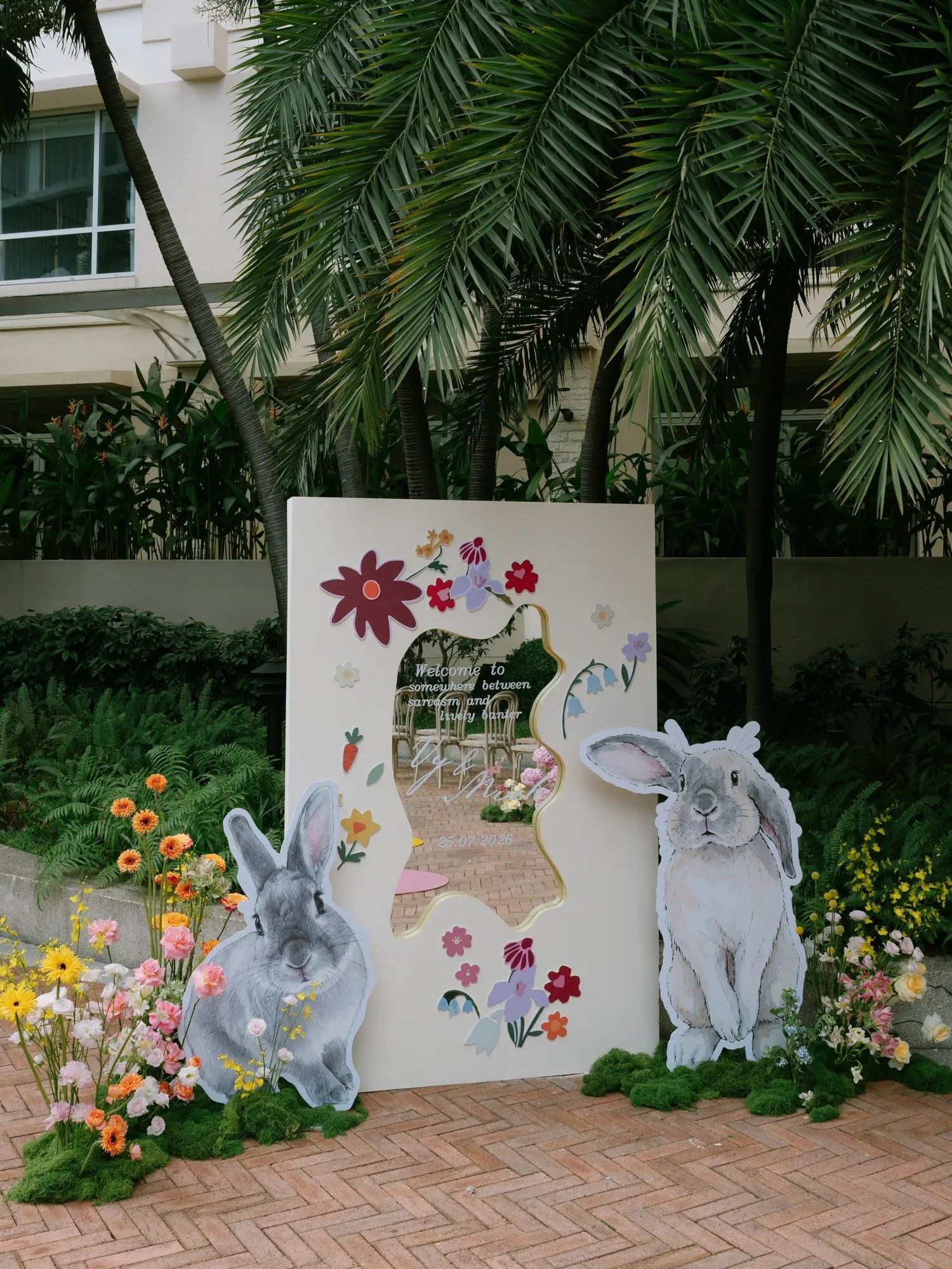Wedding welcome sign with a wavy mirror cutout reading Welcome to somewhere between sarcasm and loving banter, flanked by painted lop-eared rabbit cutouts and garden flowers