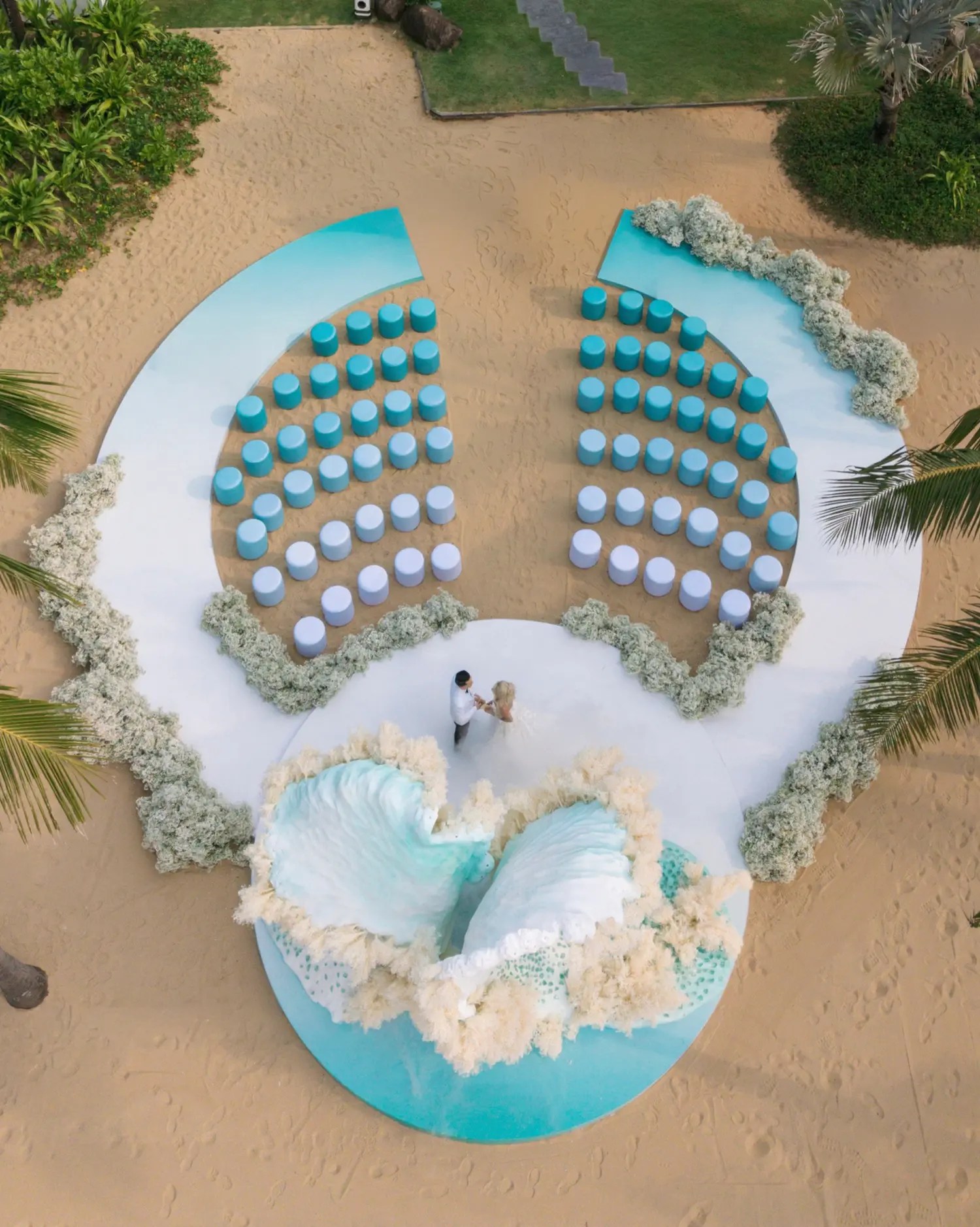 Overhead drone shot of a couple at the end of a white aisle in front of a teal wave ceremony arch, surrounded by curved rows of blue stools and cream dried flowers on the beach
