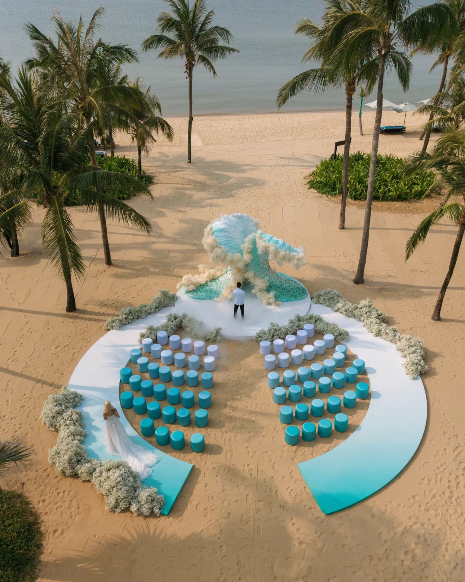 Aerial view of a beach wedding ceremony with a sculpted teal wave arch, two curved blocks of blue stools, and dried floral borders on the sand among palm trees in Danang, Vietnam