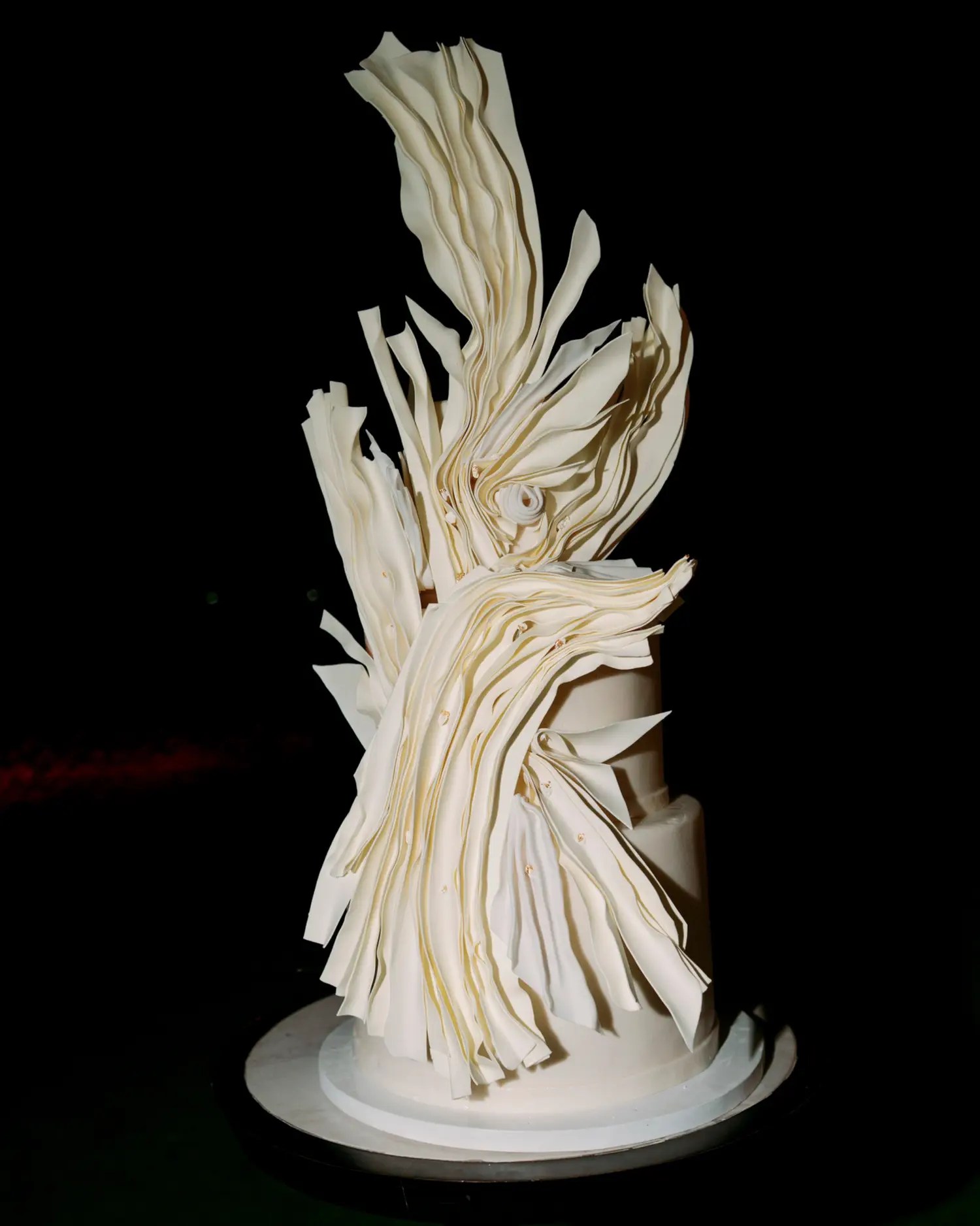 Tall white wedding cake with sculpted sugar ribbons swirling up like a breaking wave, on a black background