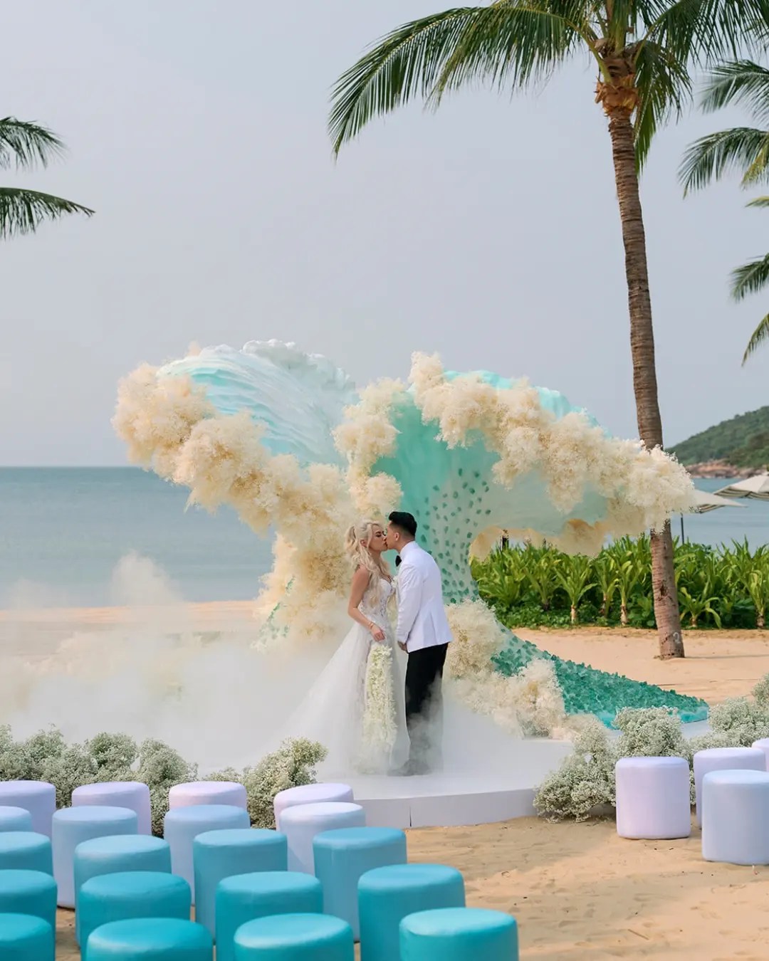 Bride and groom kissing in front of a teal wave-shaped ceremony arch with dried cream blooms on the beach at InterContinental Danang, pale blue stools in the foreground