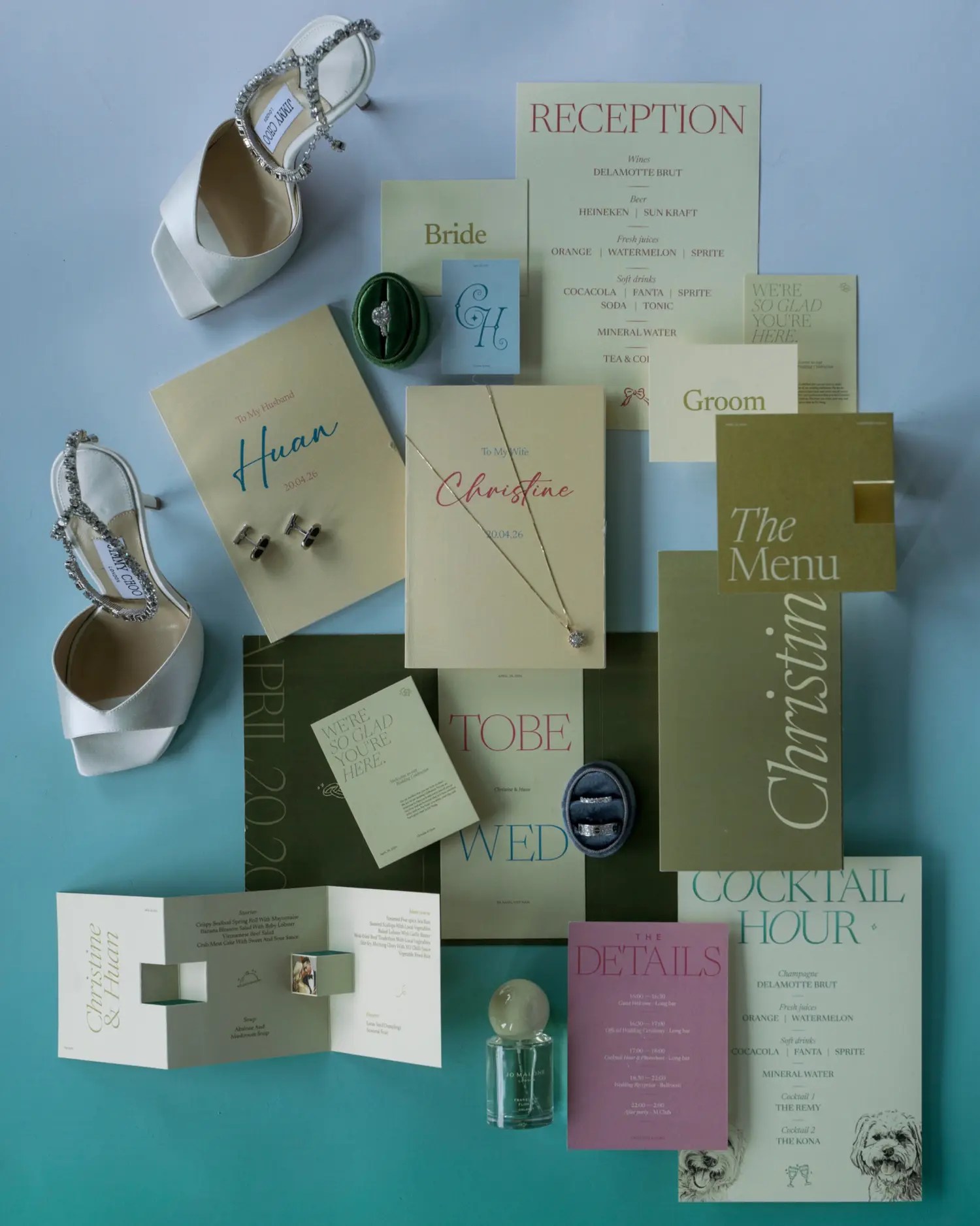 Flat lay of a modern wedding stationery suite in sage, cream, and olive with Christine and Huan in script, a cocktail hour card, a menu, a ring box, and white crystal-strap heels on a teal background