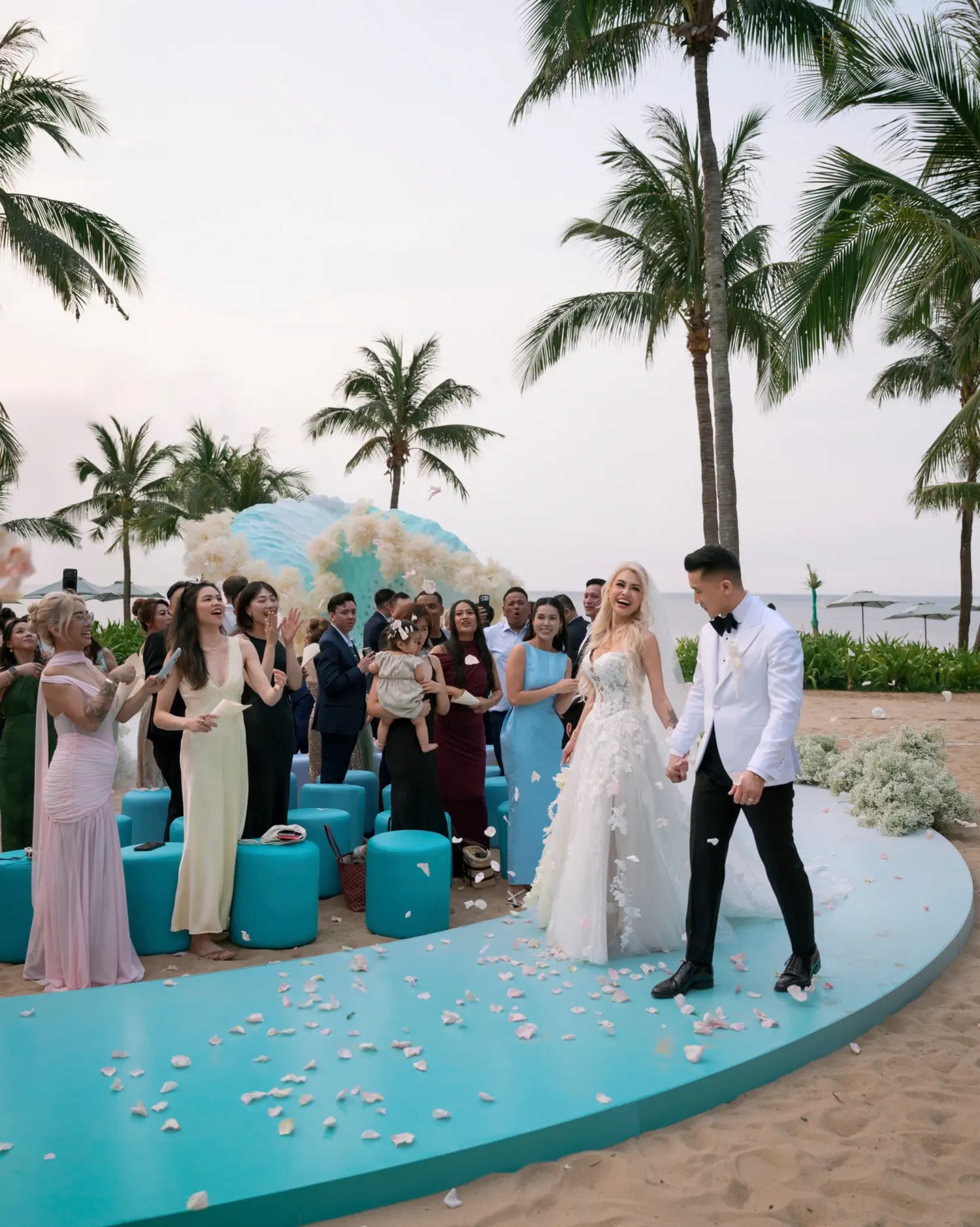Bride in a lace ballgown and groom in a white dinner jacket walking up a petal-strewn teal aisle as guests cheer beside blue stools and palm trees