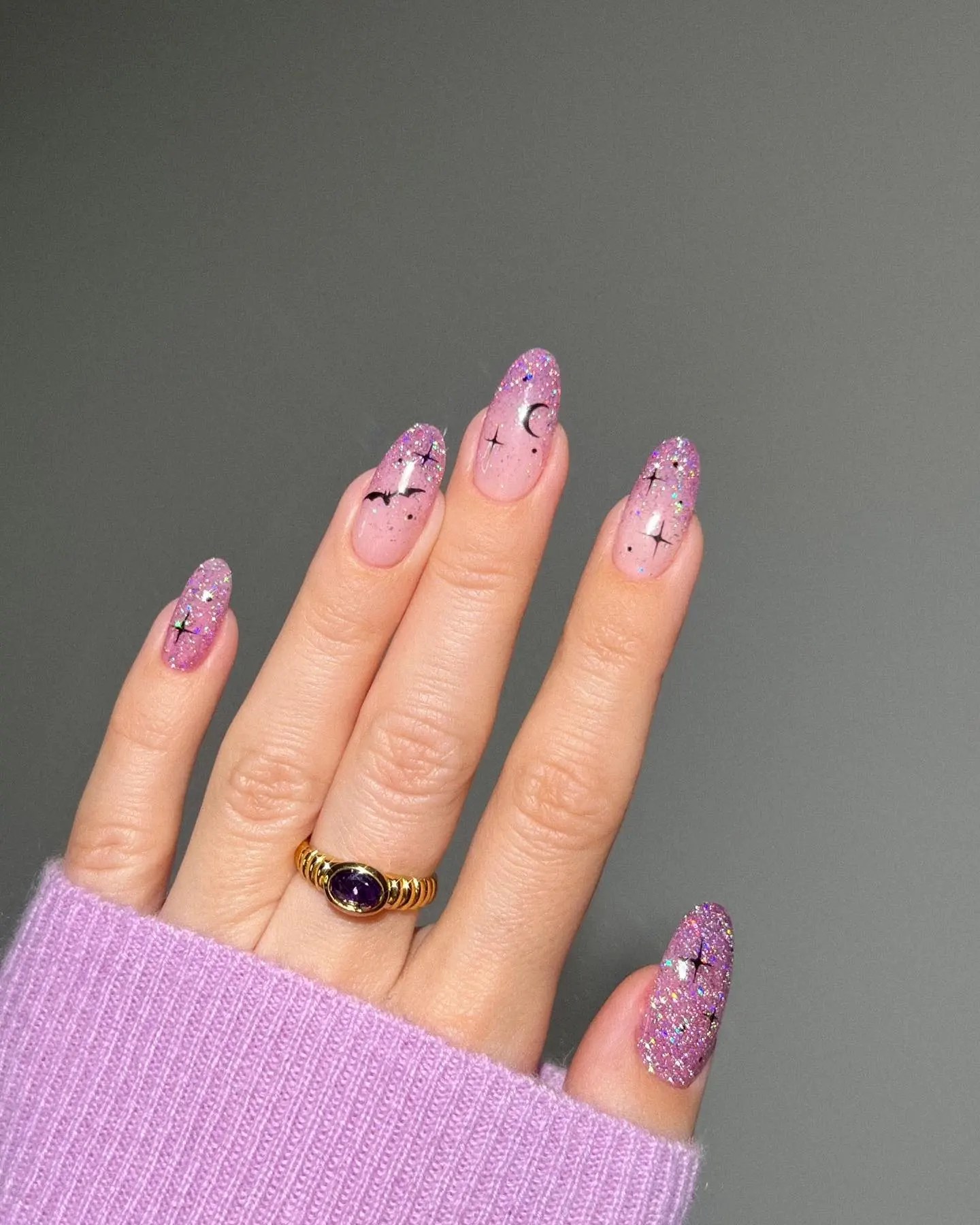 Long almond nails in a sheer lilac with holographic glitter, accented with tiny black bats, a crescent moon and small stars, worn with a gold amethyst ring and a lilac sweater.
