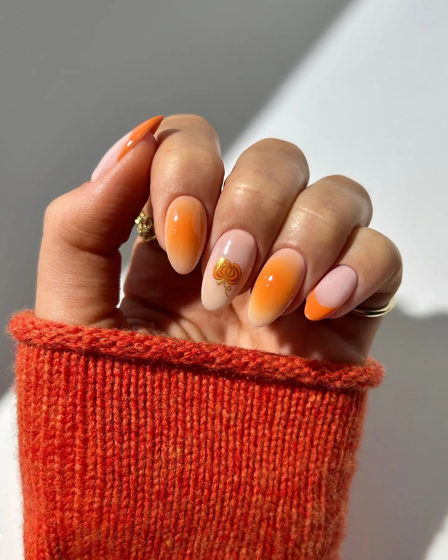 Almond nails with orange aura gradients, orange French tips and one nude accent nail with a small gold pumpkin decal, held against a burnt orange knit sweater.