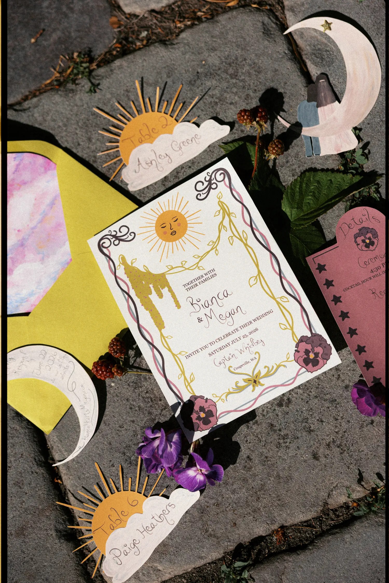 Whimsical wedding invitation suite flat lay with a sun-face illustrated invitation, cloud-shaped escort cards, a crescent moon RSVP card, mustard envelopes, and loose blackberries