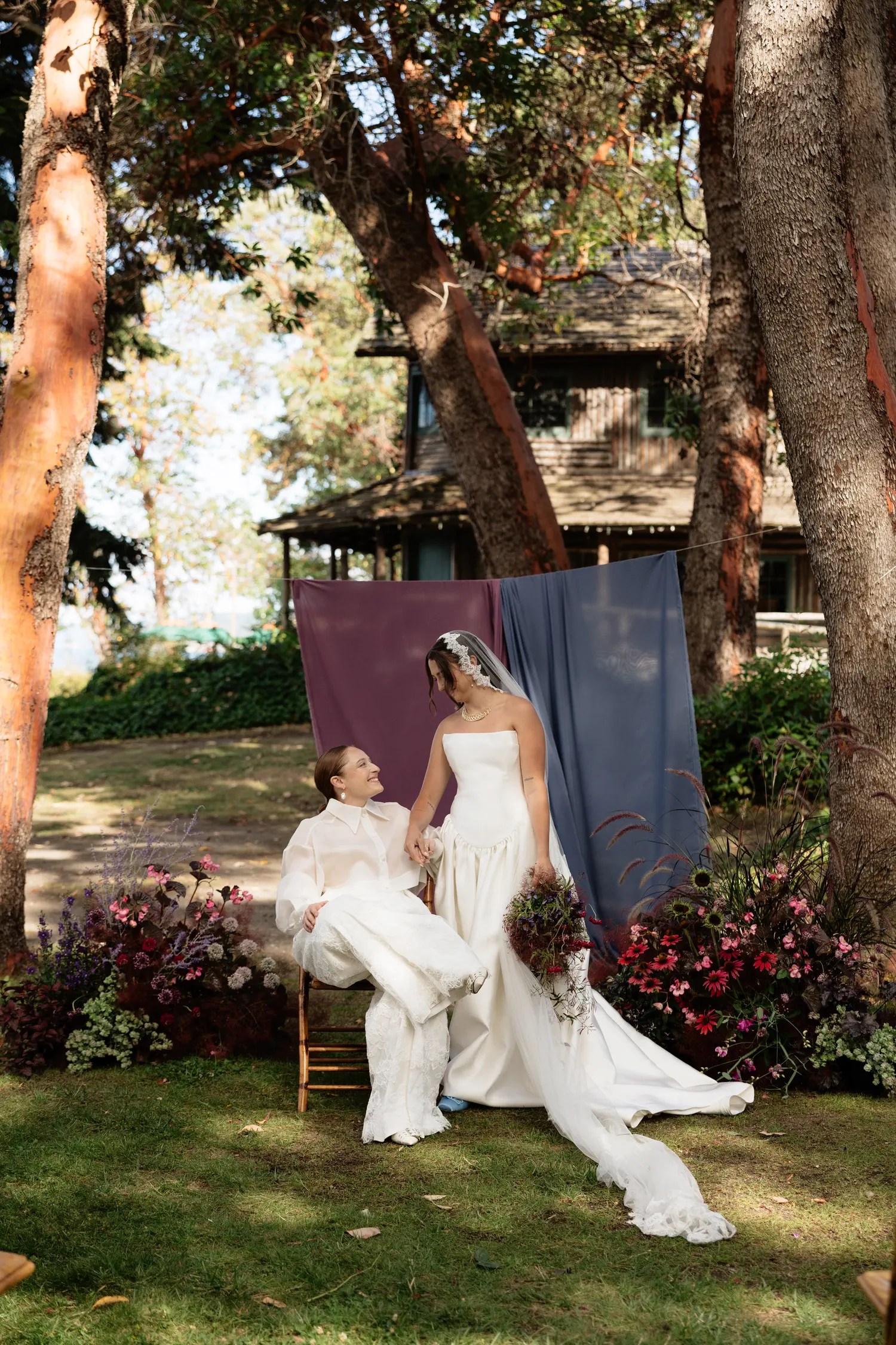 Bride in an organza barong seated on a chair looking up at her bride in a satin gown and veil, framed by madrona trees and plum and dusty blue chiffon drapes