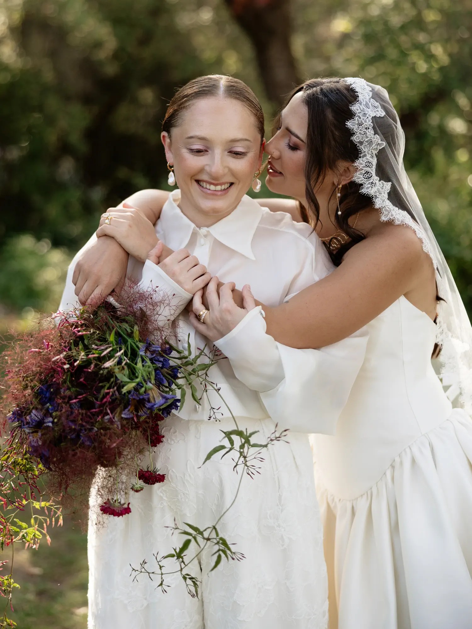 Two brides embracing, one in an organza barong and one in a satin gown with lace veil, both in gold hoop earrings and holding a wildflower bouquet