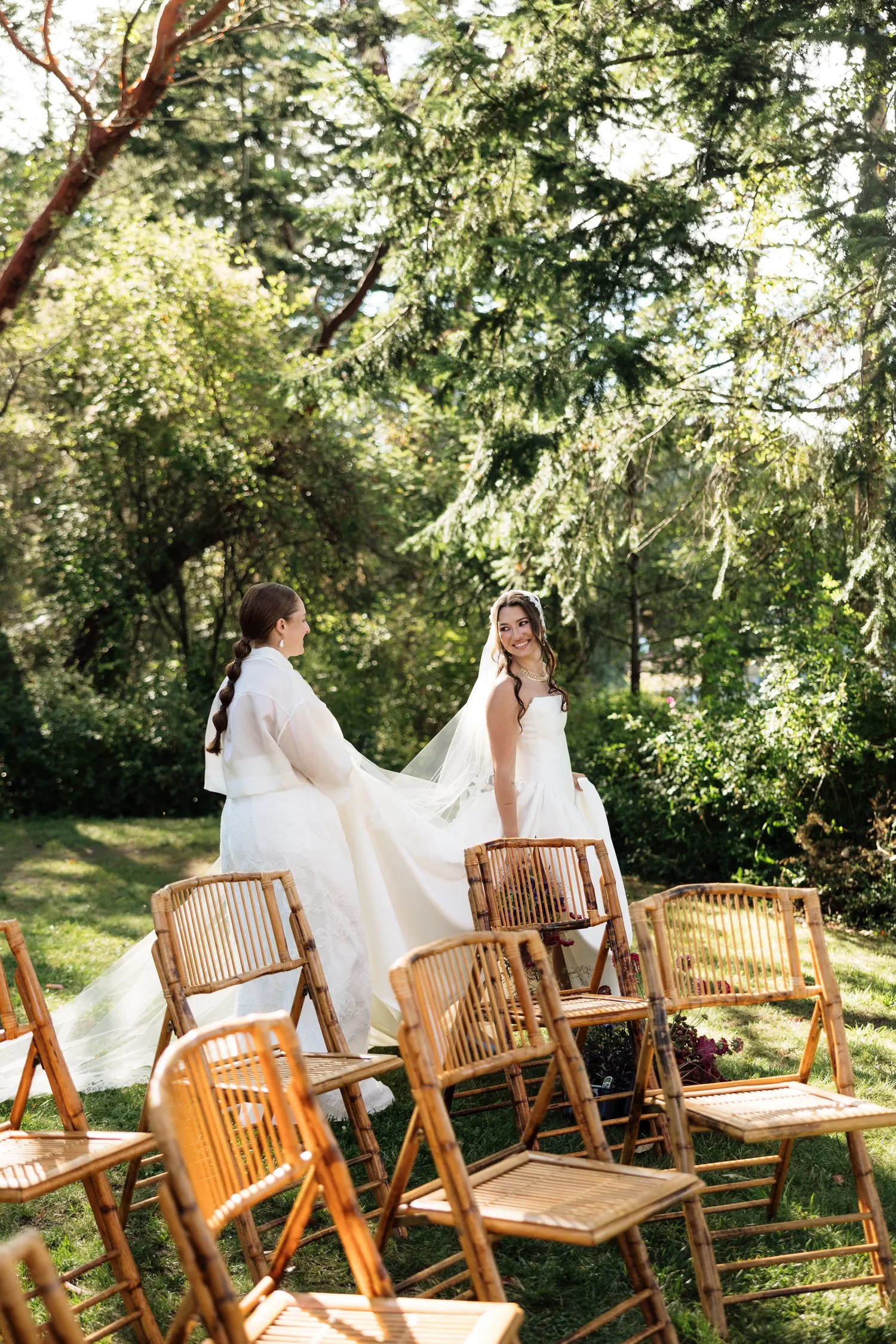 Two brides at the end of a bamboo chair-lined ceremony aisle under evergreen trees, one lifting the train of the other's satin gown