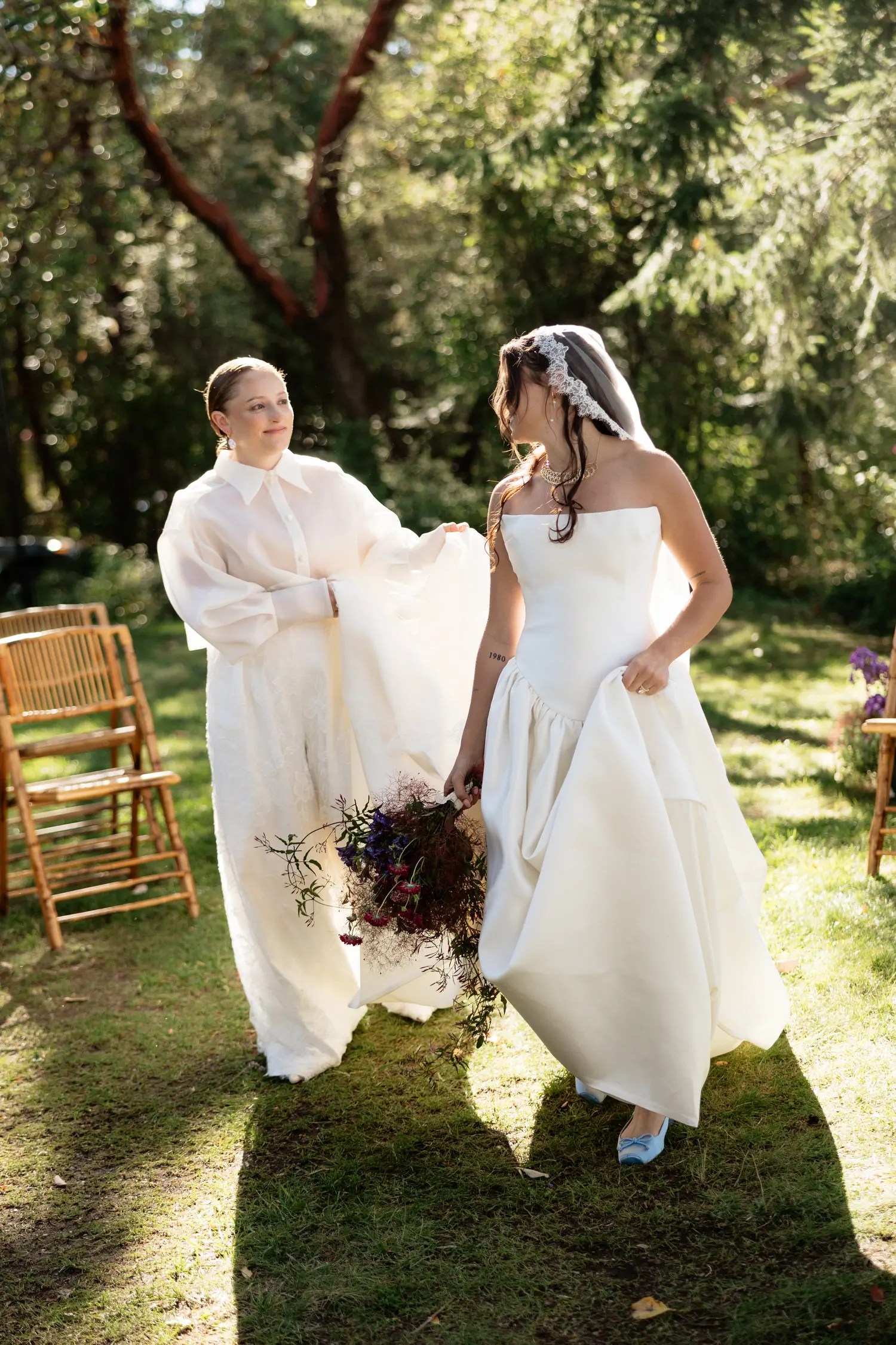 Bride in an organza barong and lace trousers carrying the ivory satin train of her bride's gown down a grass aisle lined with bamboo chairs