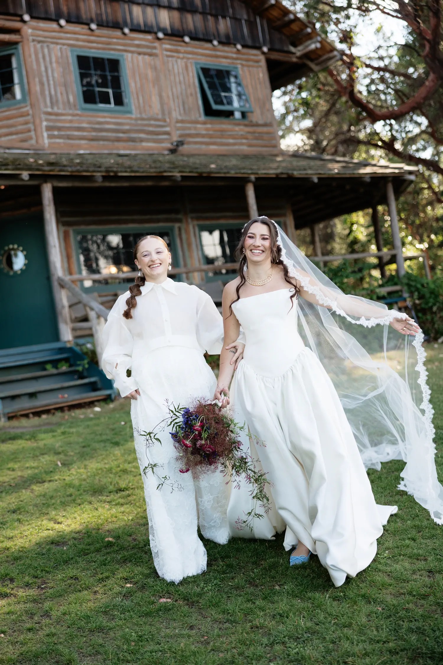 Two laughing brides walking in front of the teal-trimmed wooden lodge at Captain Whidbey, lace veil blowing in the wind and light blue flats under the gown