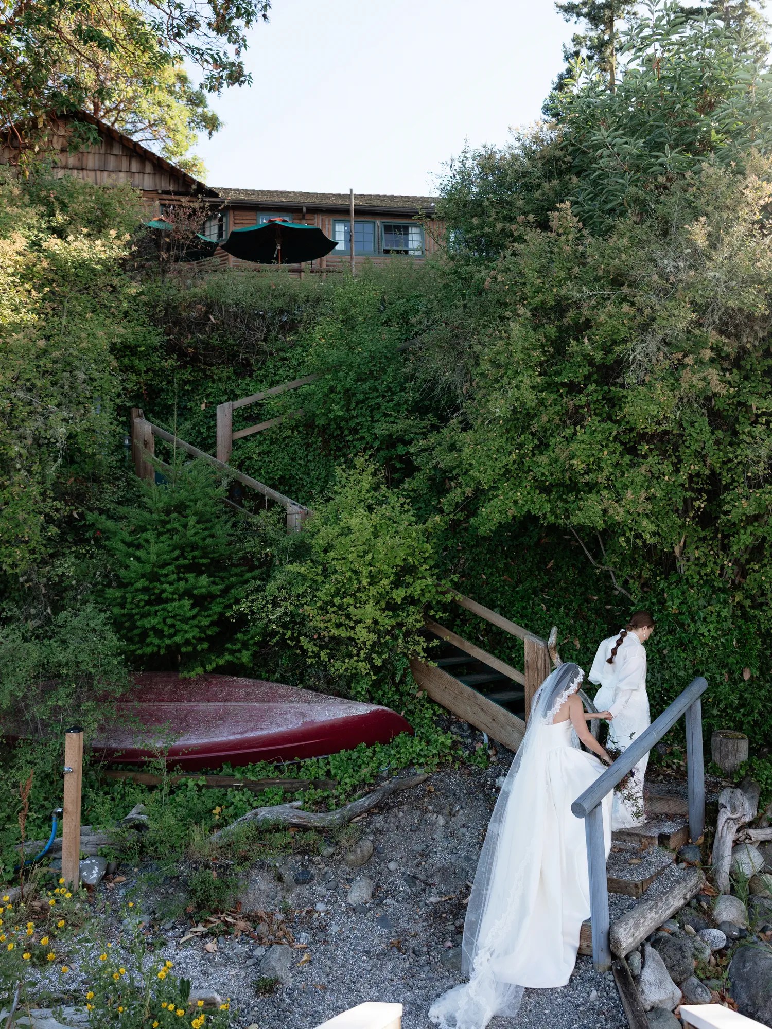 Two brides in white climbing a wooden staircase through greenery toward a cedar-shingled building at Captain Whidbey, with a red canoe on the gravel below