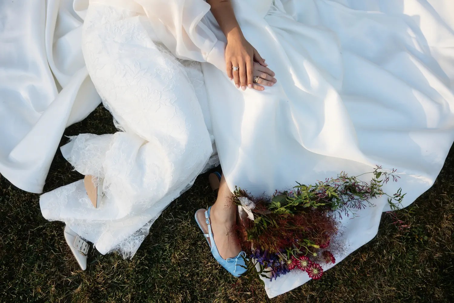 Overhead detail of two brides' hands with gold rings, a light blue slingback flat with a bow, and a wildflower bouquet resting on an ivory satin skirt