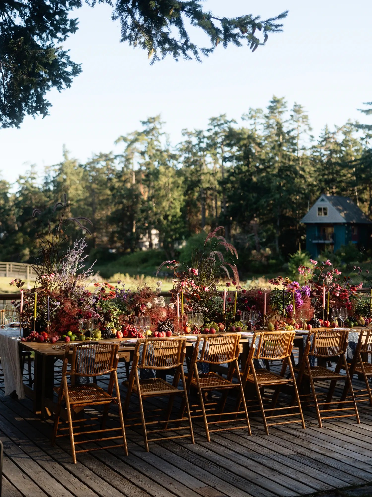 Long communal wedding reception table on a wooden deck at Captain Whidbey with a wild fall floral and fruit centerpiece, taper candles, bamboo chairs, and a teal A-frame cottage behind