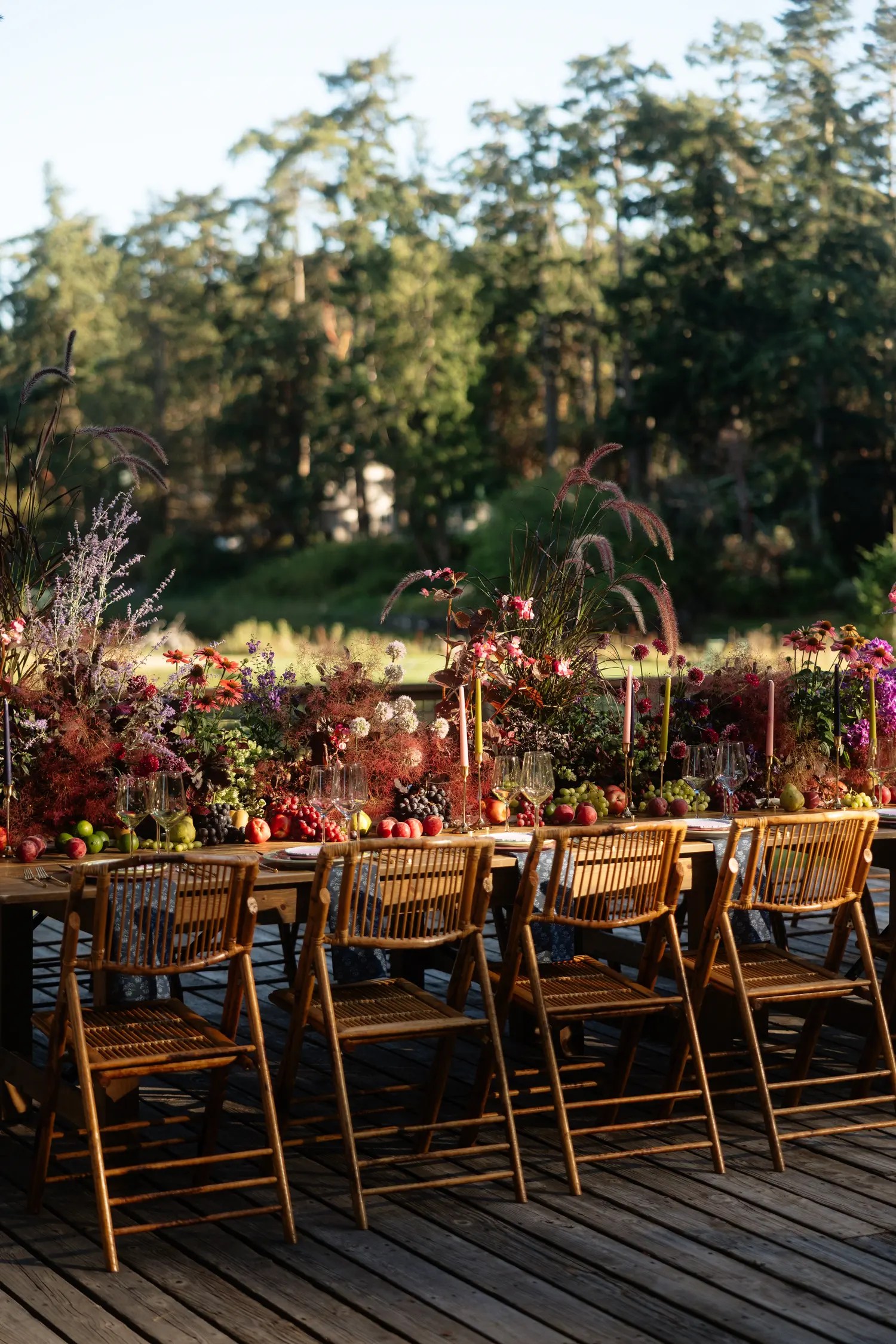 Fall wedding table centerpiece of burgundy ornamental grasses, purple dahlias, lavender blooms, apples, pears, and grapes with mauve and green taper candles on an outdoor deck