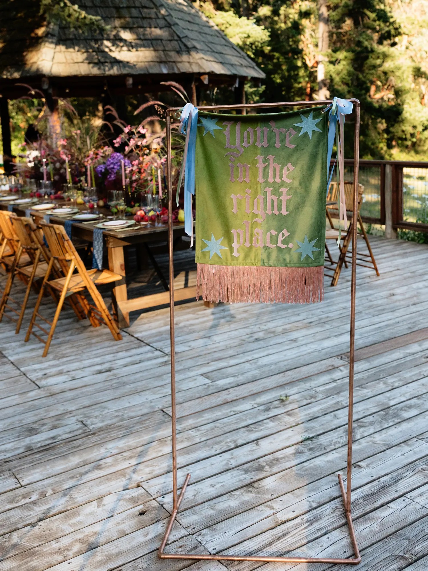 Olive green wedding welcome banner reading You're in the right place with dusty pink fringe and blue stars hanging from a copper garment rack beside a reception table