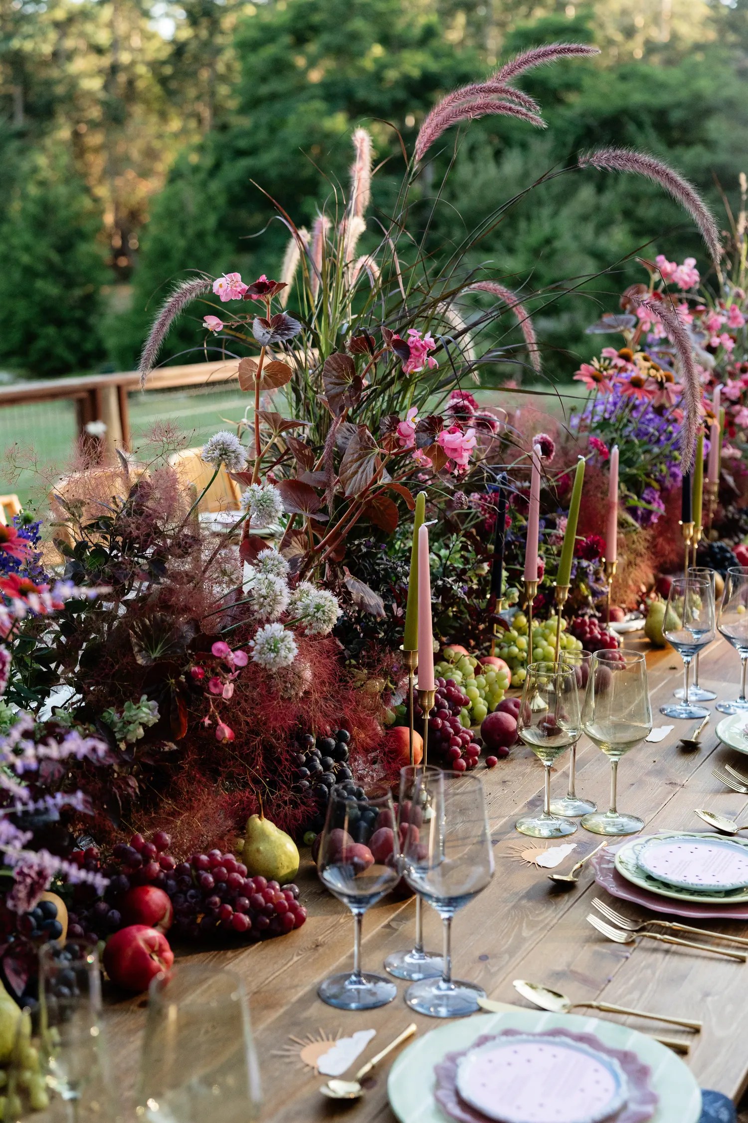 Corner of a fall wedding tablescape with sage green and scalloped mauve charger plates, gold flatware, blue wine glasses, grapes, pears, dahlias, and arching ornamental grass plumes