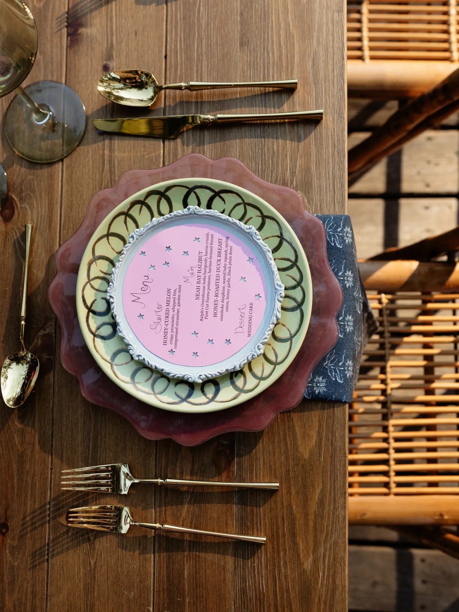 Overhead wedding place setting with a round pink star-dotted menu card listing starter, main, and dessert on layered mauve and sage plates with gold flatware