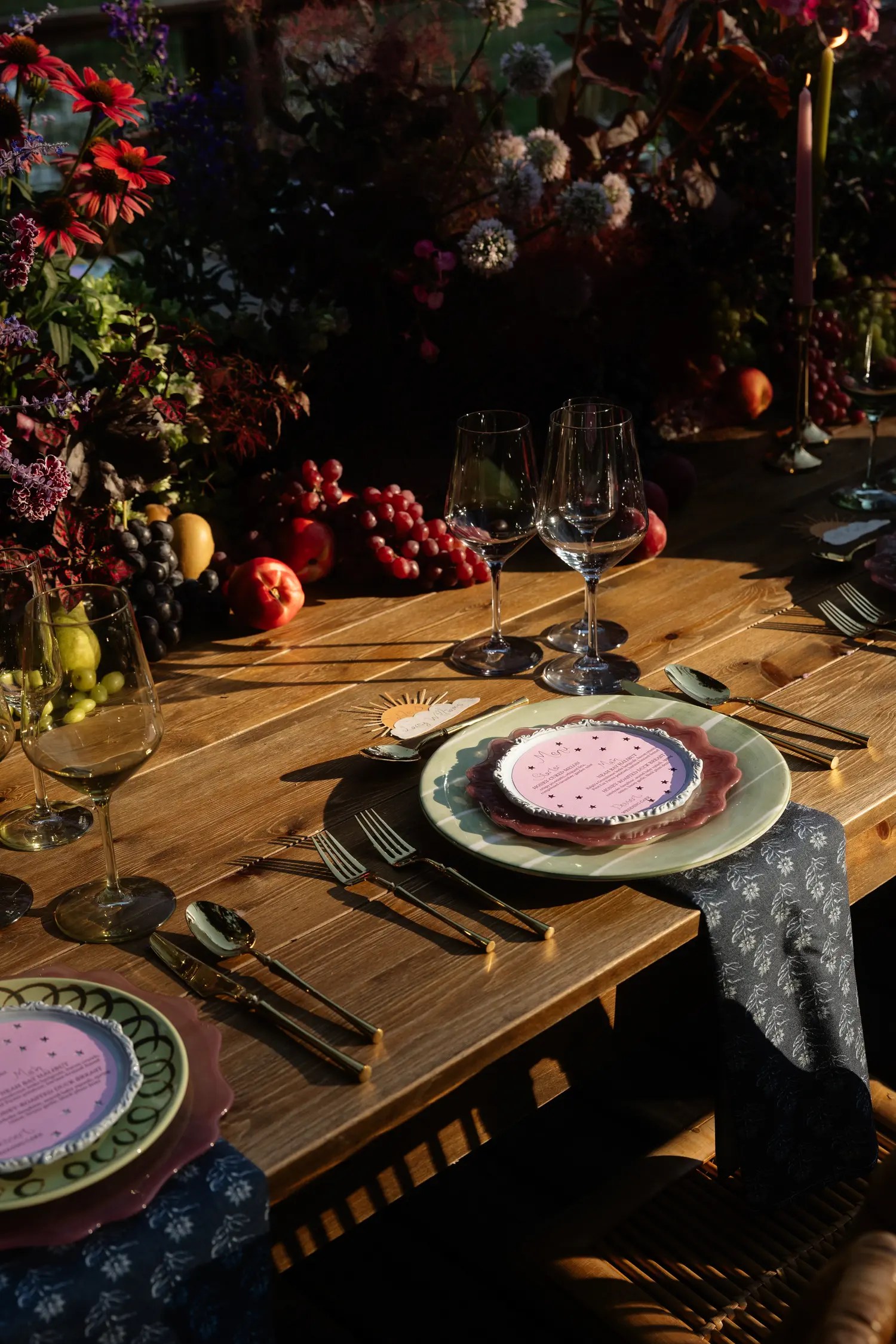 Moody side-lit wedding table with stacked sage and mauve chargers, dark gold flatware, and dark grapes, apples, and plums tucked into a burgundy and purple floral centerpiece