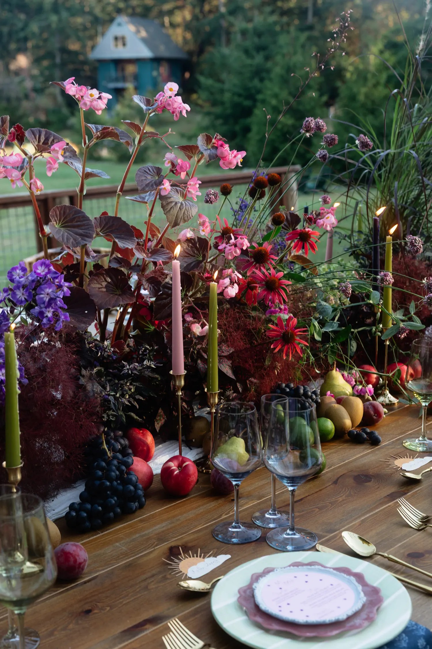 Lit pink taper candle in a brass candlestick among dark maroon begonia leaves, coneflowers, smoke bush, grapes, and plums on a dusk wedding table