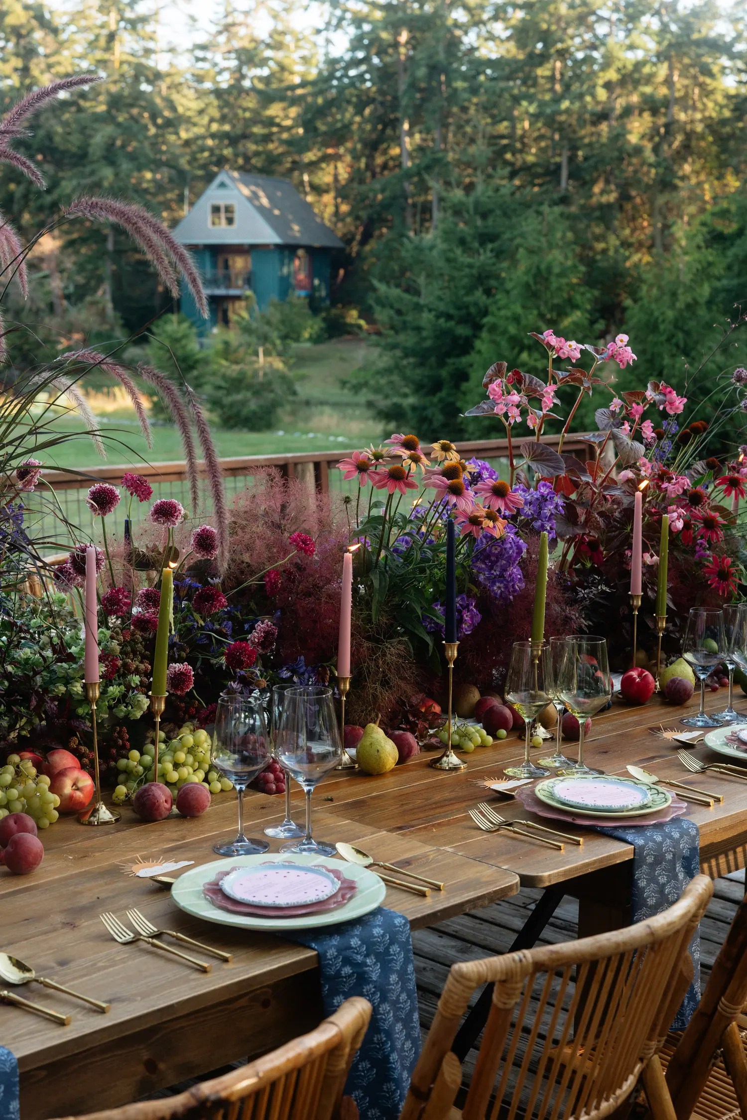 Fall wedding reception table at dusk with lit olive, pink, and plum taper candles, coneflowers, dahlias, scattered fruit, and a teal cottage beyond the deck railing