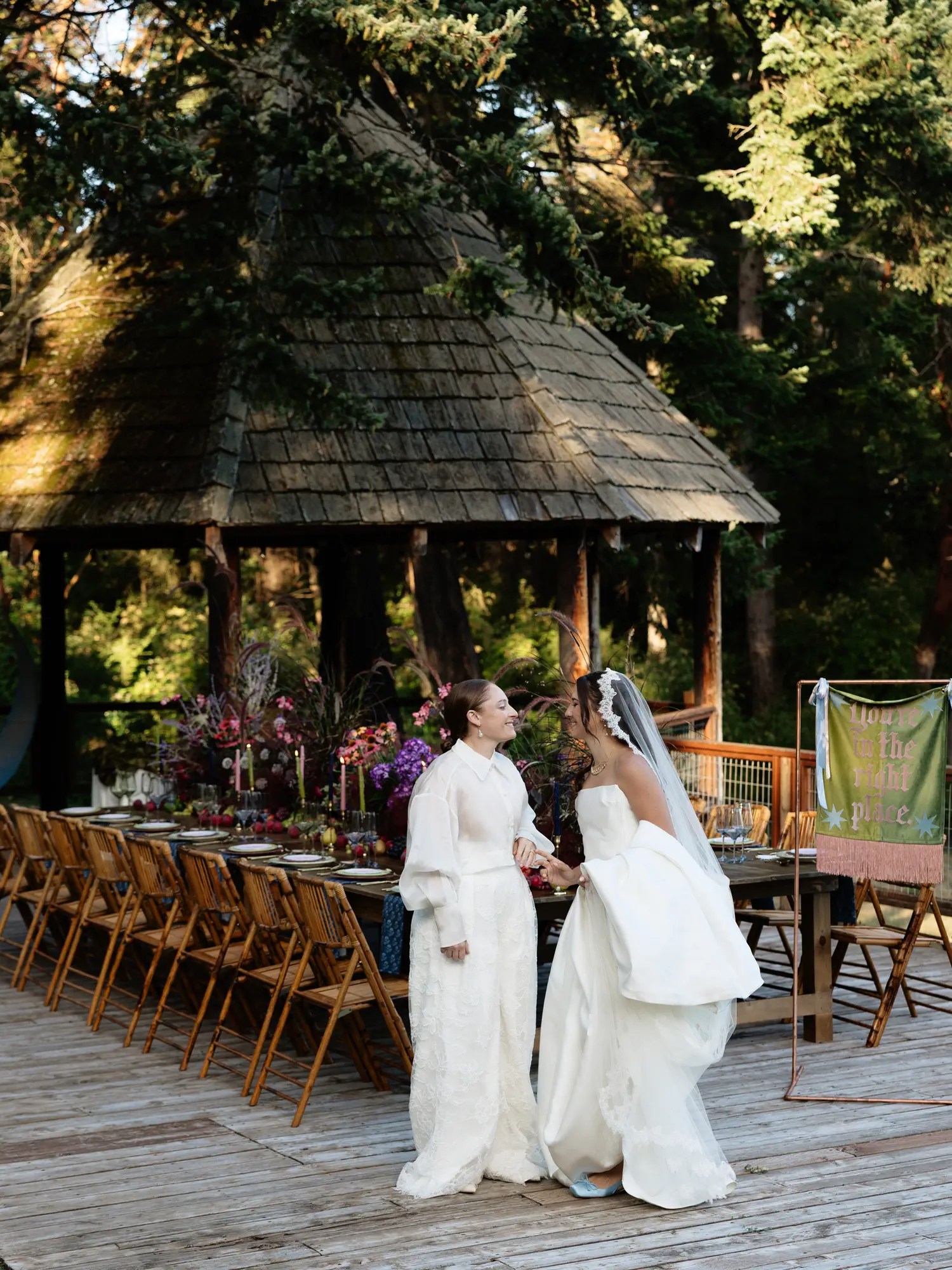 Two brides in reception looks, a boxy white jacket with wide trousers and a strapless gown with an oversized hip bow and floral veil, holding hands on a deck beside a long reception table and gazebo