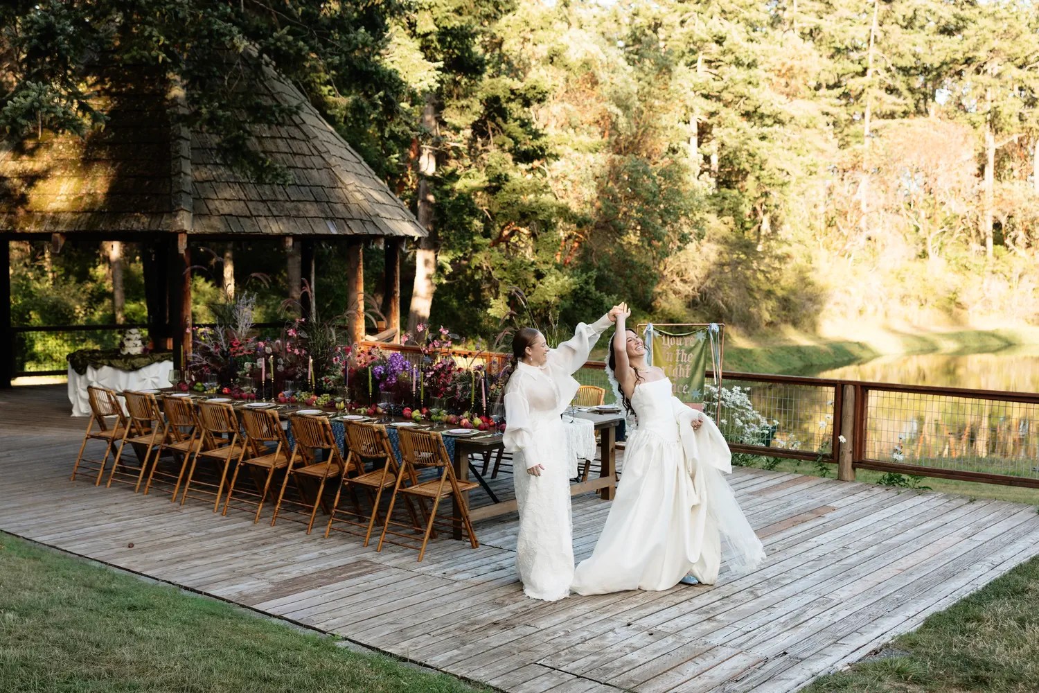 Two brides dancing on a wooden deck beside a long floral reception table with a You're in the right place banner, a gazebo, and the lagoon at Captain Whidbey behind