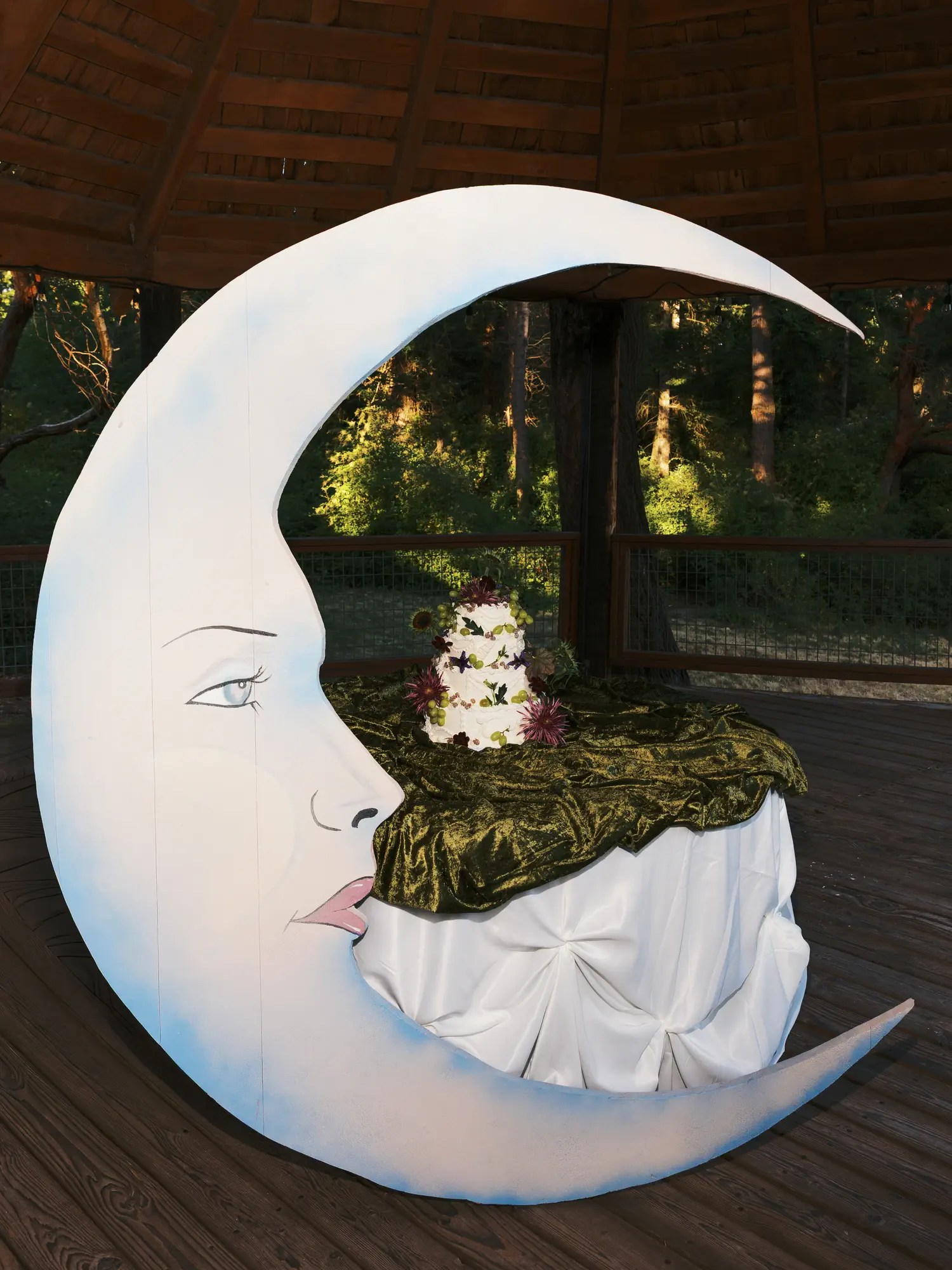 Large white crescent moon cake display prop with a painted face holding a three-tier white wedding cake on olive crushed velvet inside a wooden gazebo