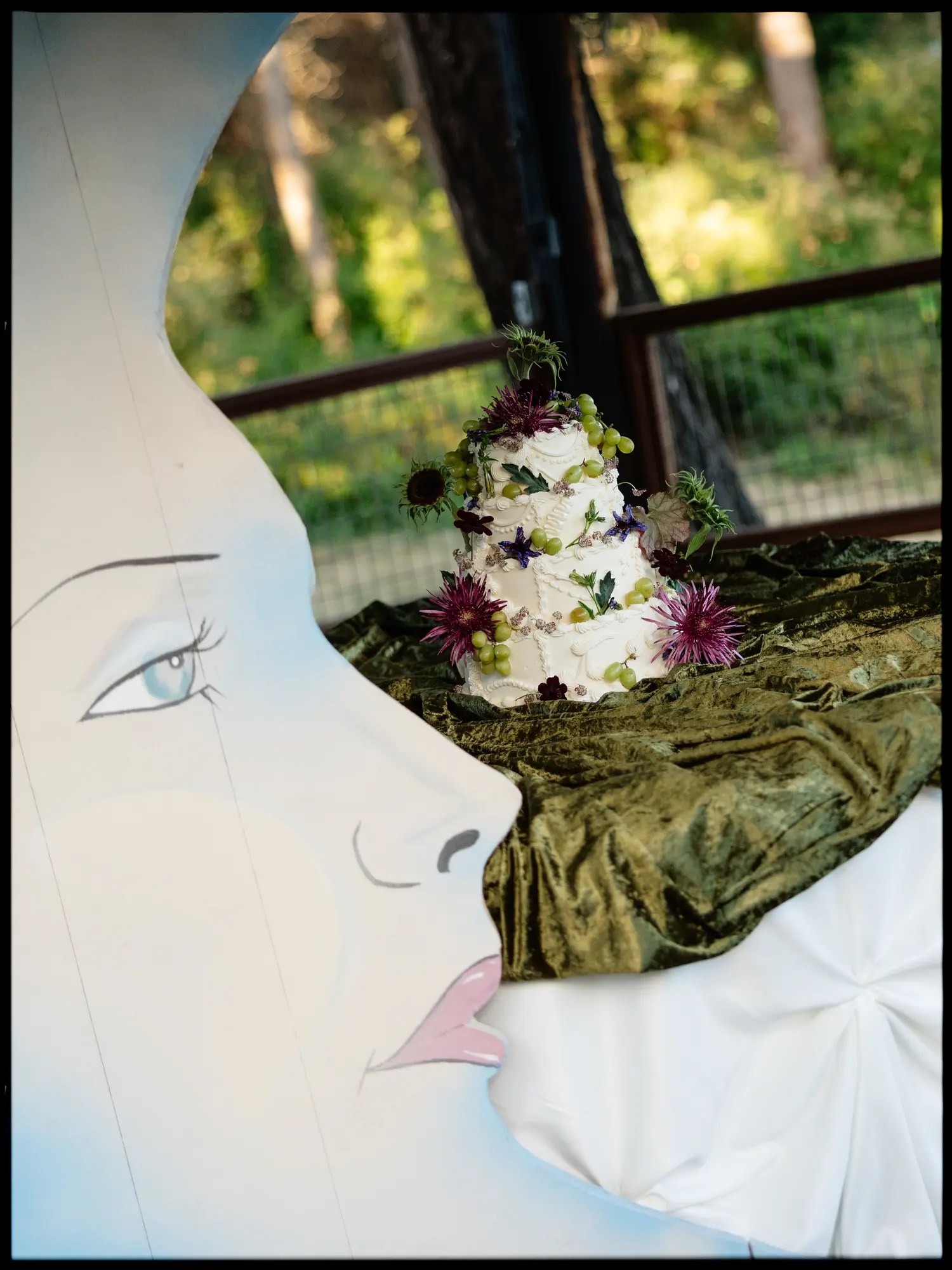 Close-up of a crescent moon cake stand prop with painted blue eyes and pink lips, with a three-tier white wedding cake decorated with purple chrysanthemums and green grapes