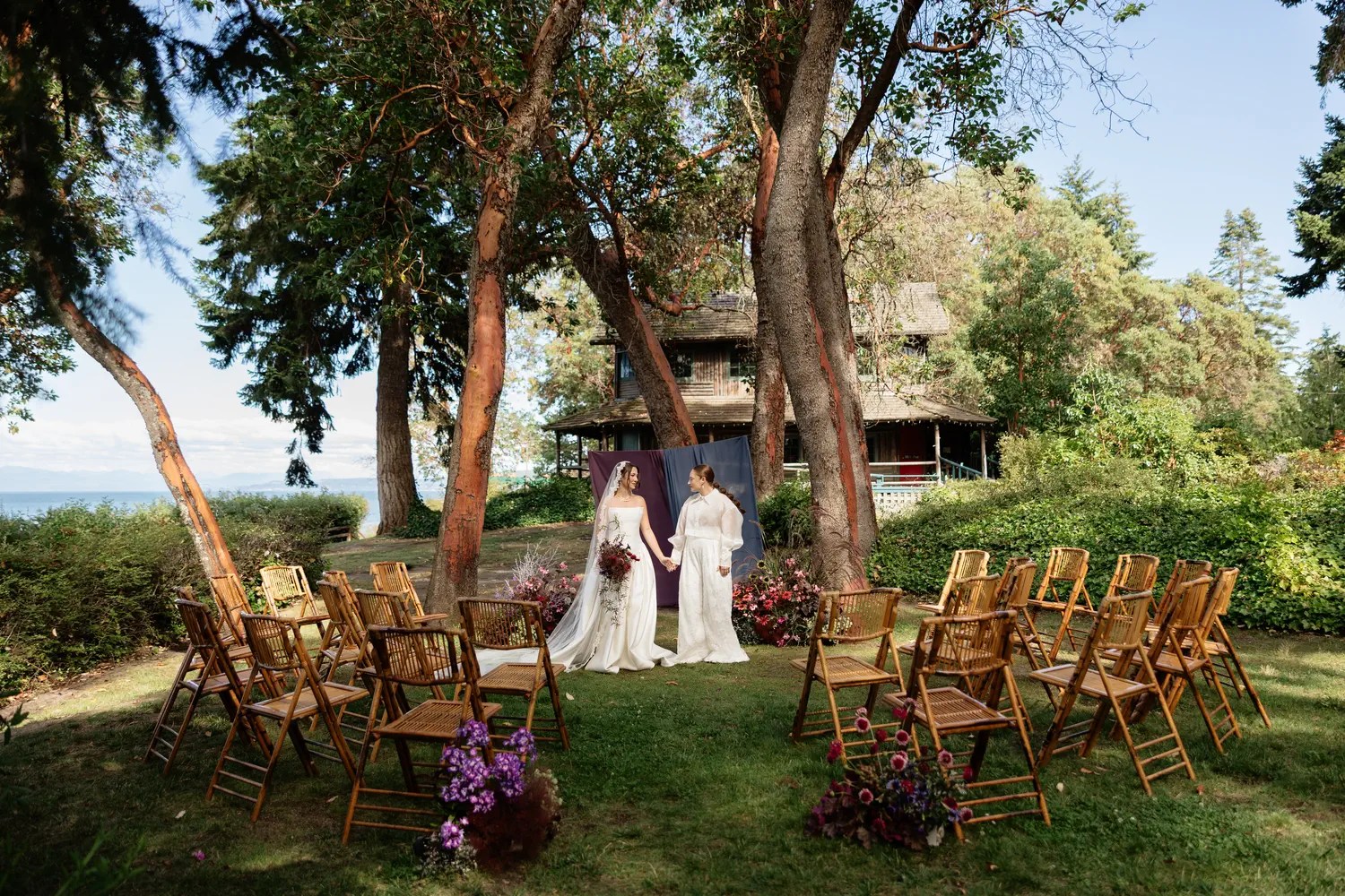 Outdoor wedding ceremony in the round at Captain Whidbey with bamboo folding chairs in a semicircle under madrona trees and a plum and dusty blue chiffon backdrop