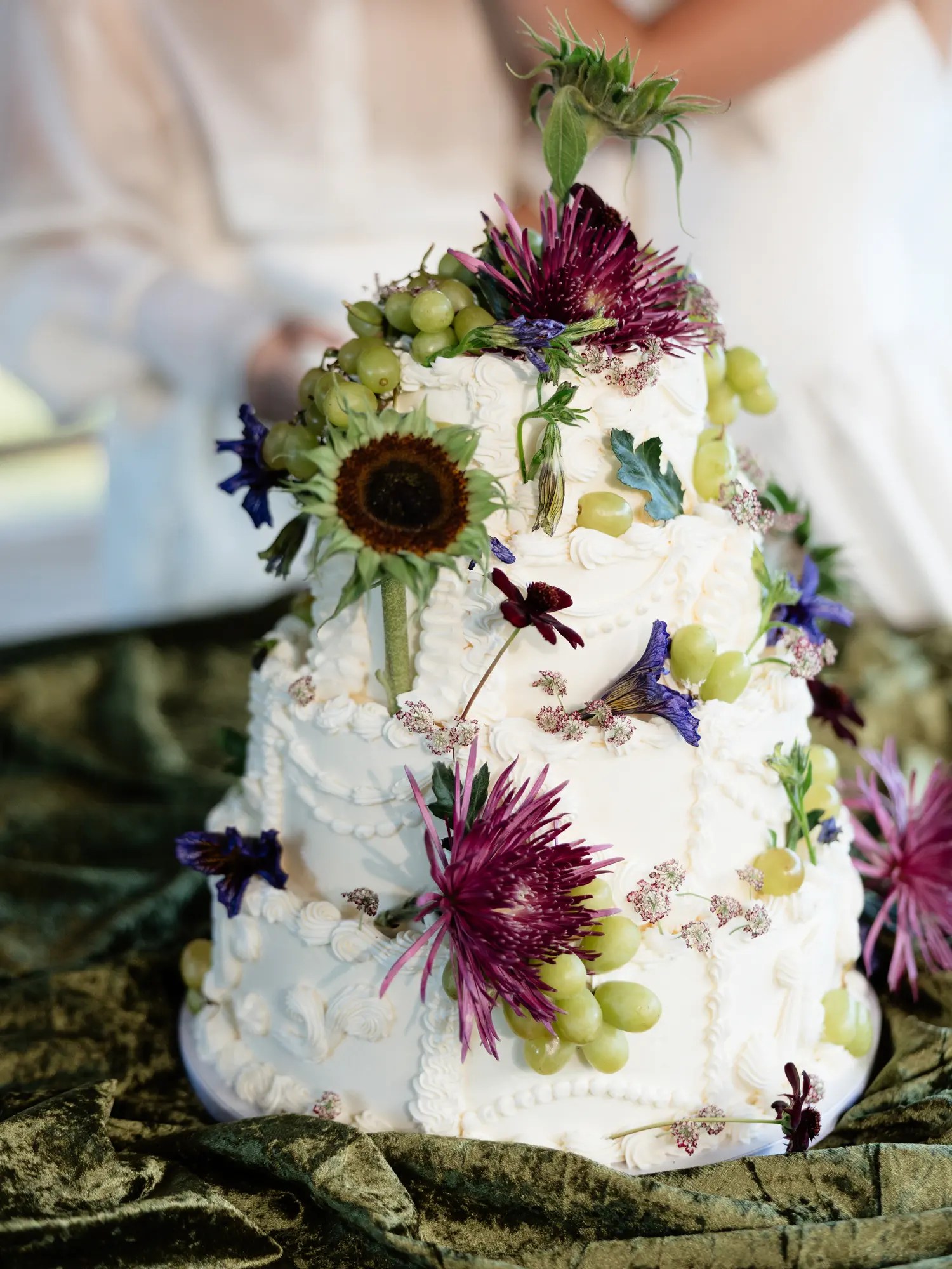 Three-tier white wedding cake with vintage piped scrolls and ruffles decorated with spiky purple chrysanthemums, green grapes, a sunflower, blue delphinium petals, and a thistle on top