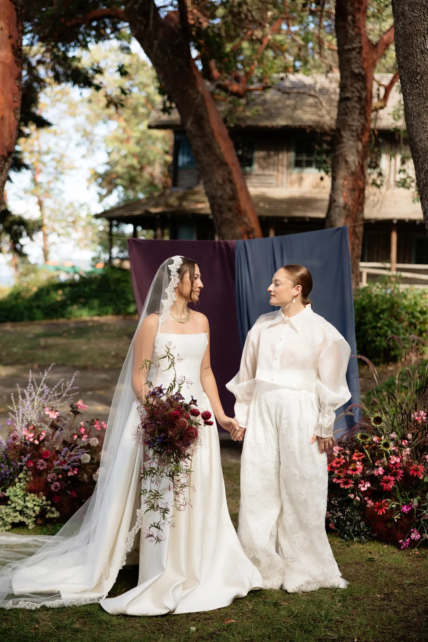 Two brides holding hands under madrona trees at Captain Whidbey, one in a strapless satin gown with lace-edged veil and the other in a sheer organza barong with wide-leg lace trousers, in front of a plum and dusty blue chiffon ceremony backdrop