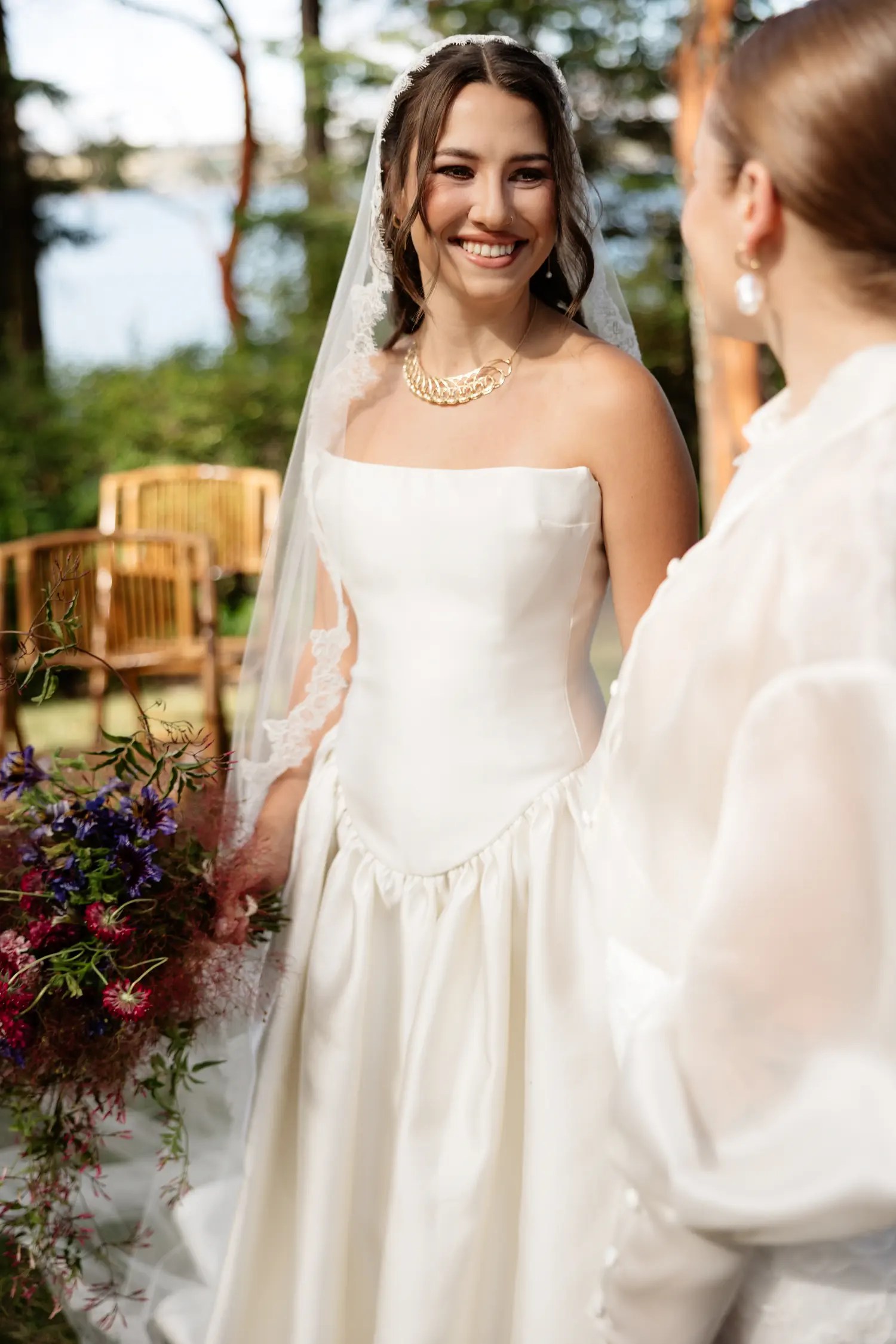 Bride in a strapless ivory satin gown, lace-edged veil, and layered gold necklace holding a cascading burgundy and purple wildflower bouquet
