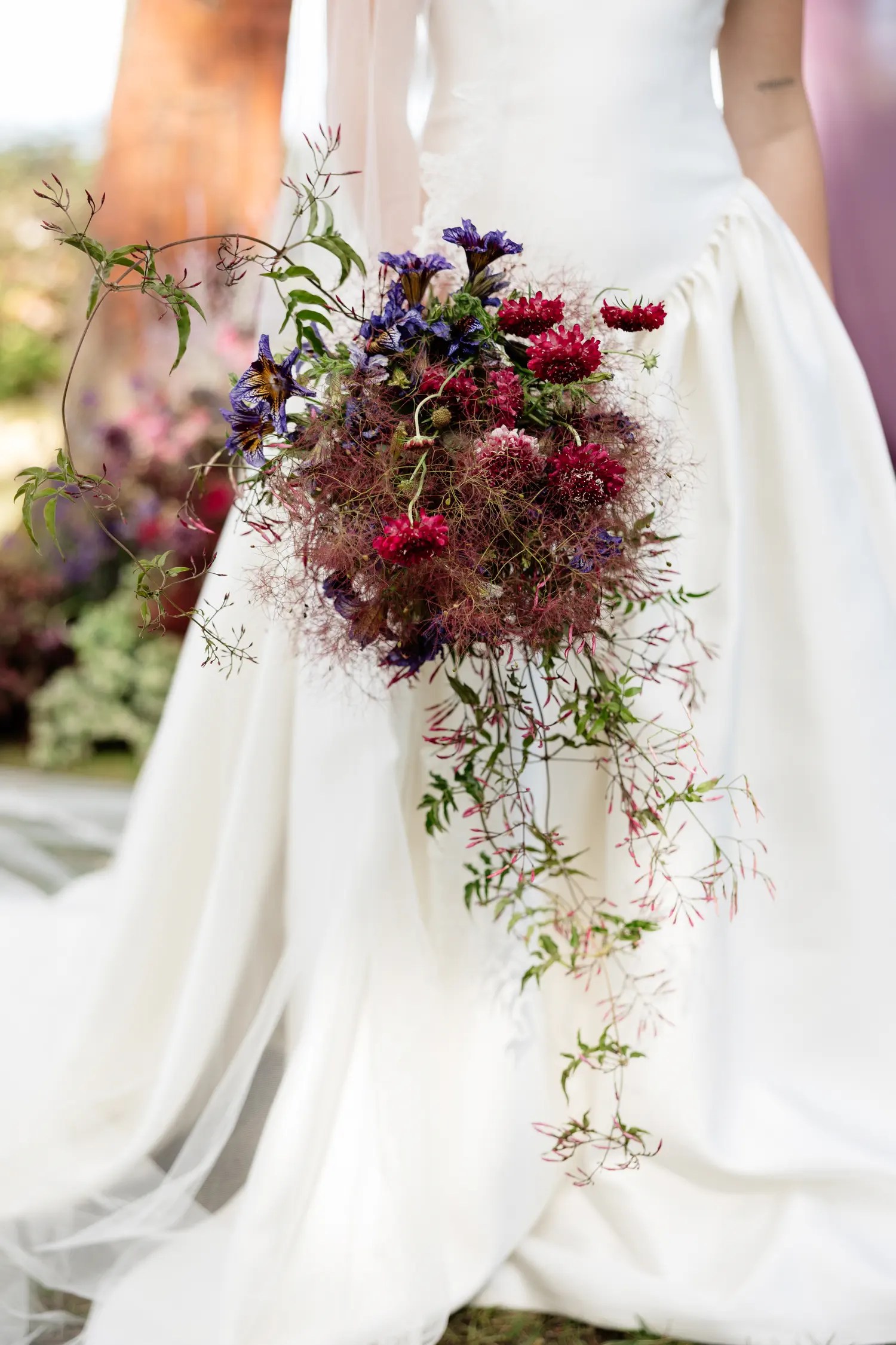 Cascading fall wedding bouquet of crimson scabiosa, purple iris, smoky mauve plumes, and trailing vine held against an ivory satin skirt
