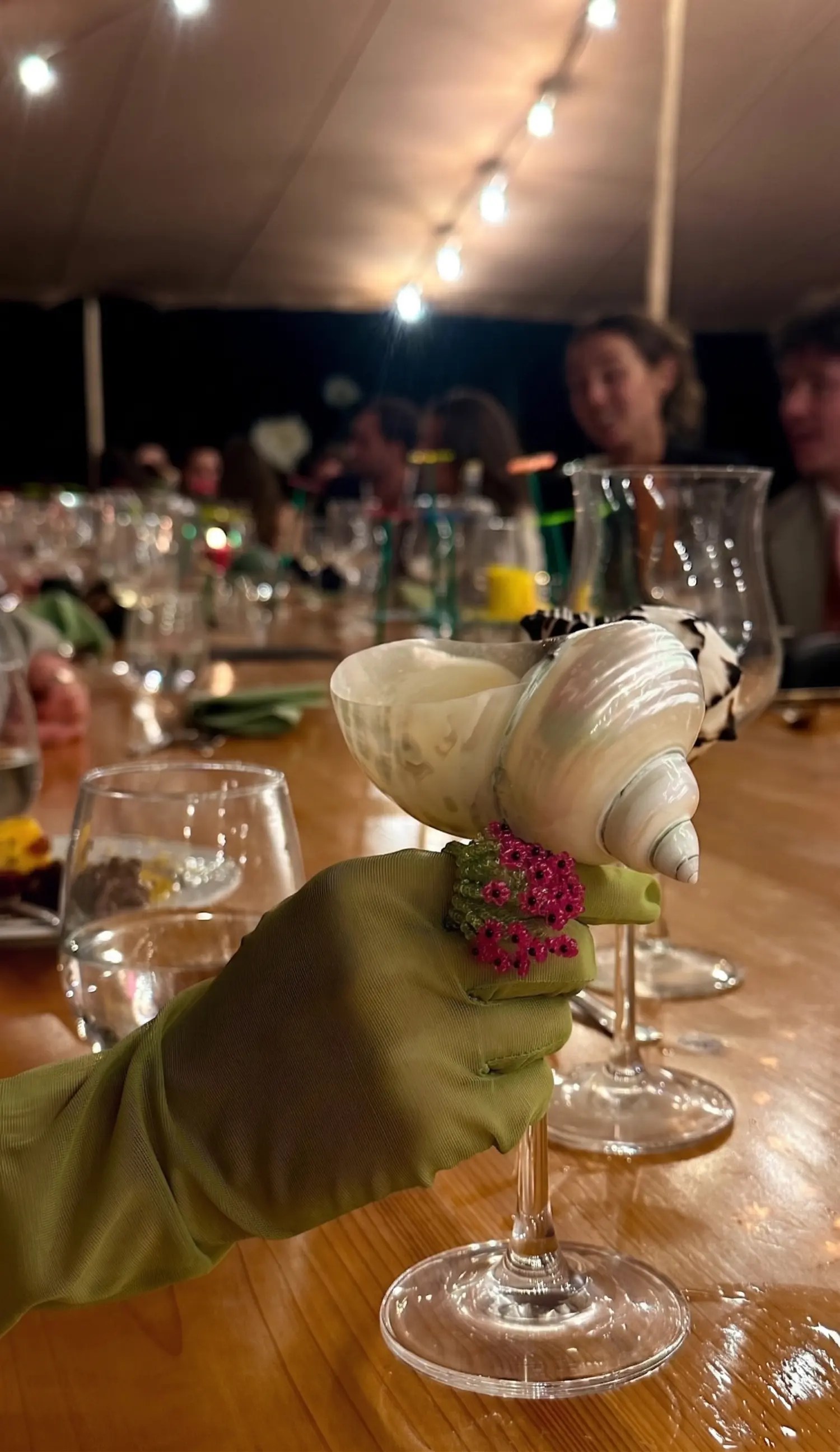 Olive green gloved hand holding a custom seashell wine glass at a candlelit tented wedding dinner in the Azores
