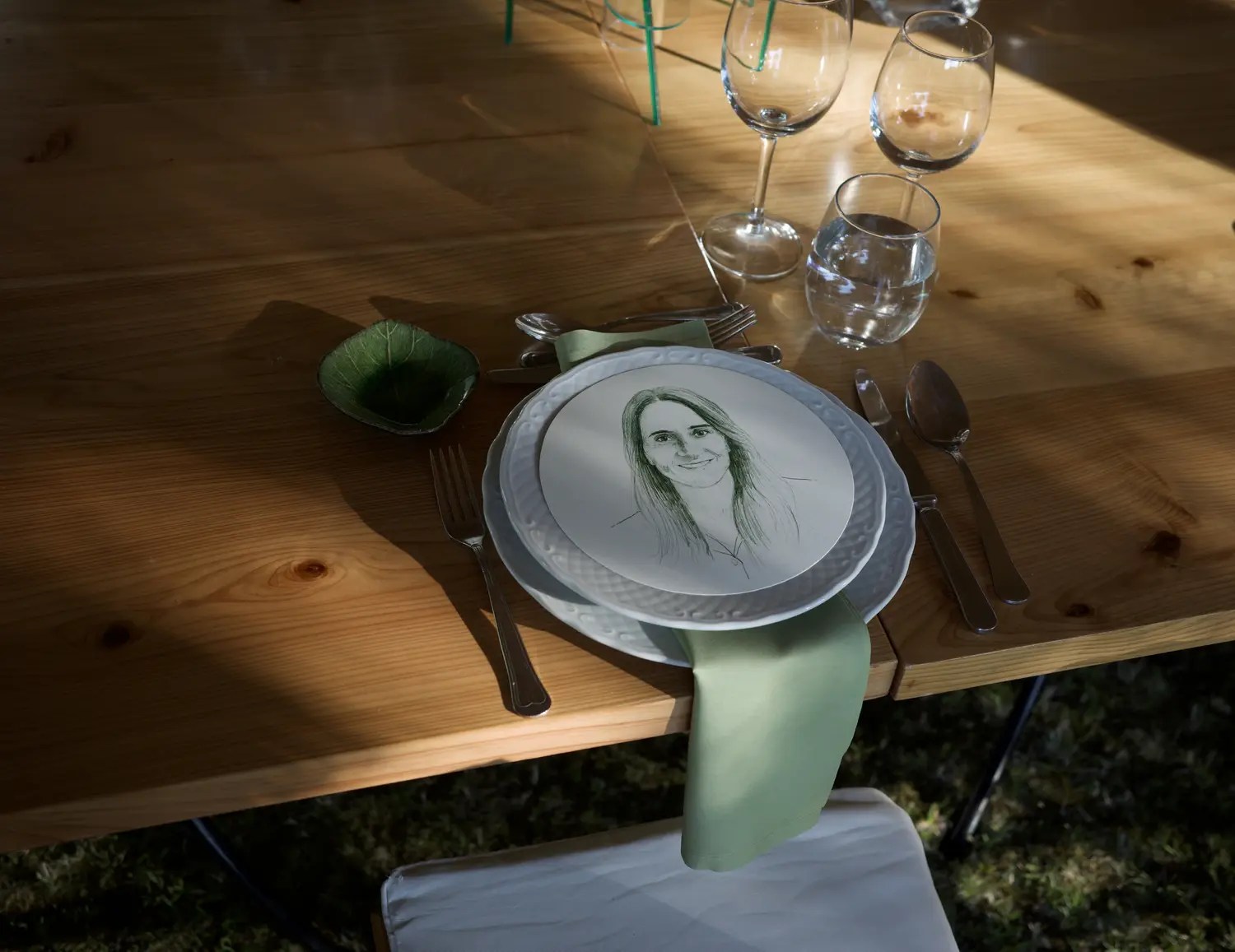 Wedding place setting on a wooden table with a hand-drawn guest portrait on a white scalloped plate, sage green napkin, and glassware