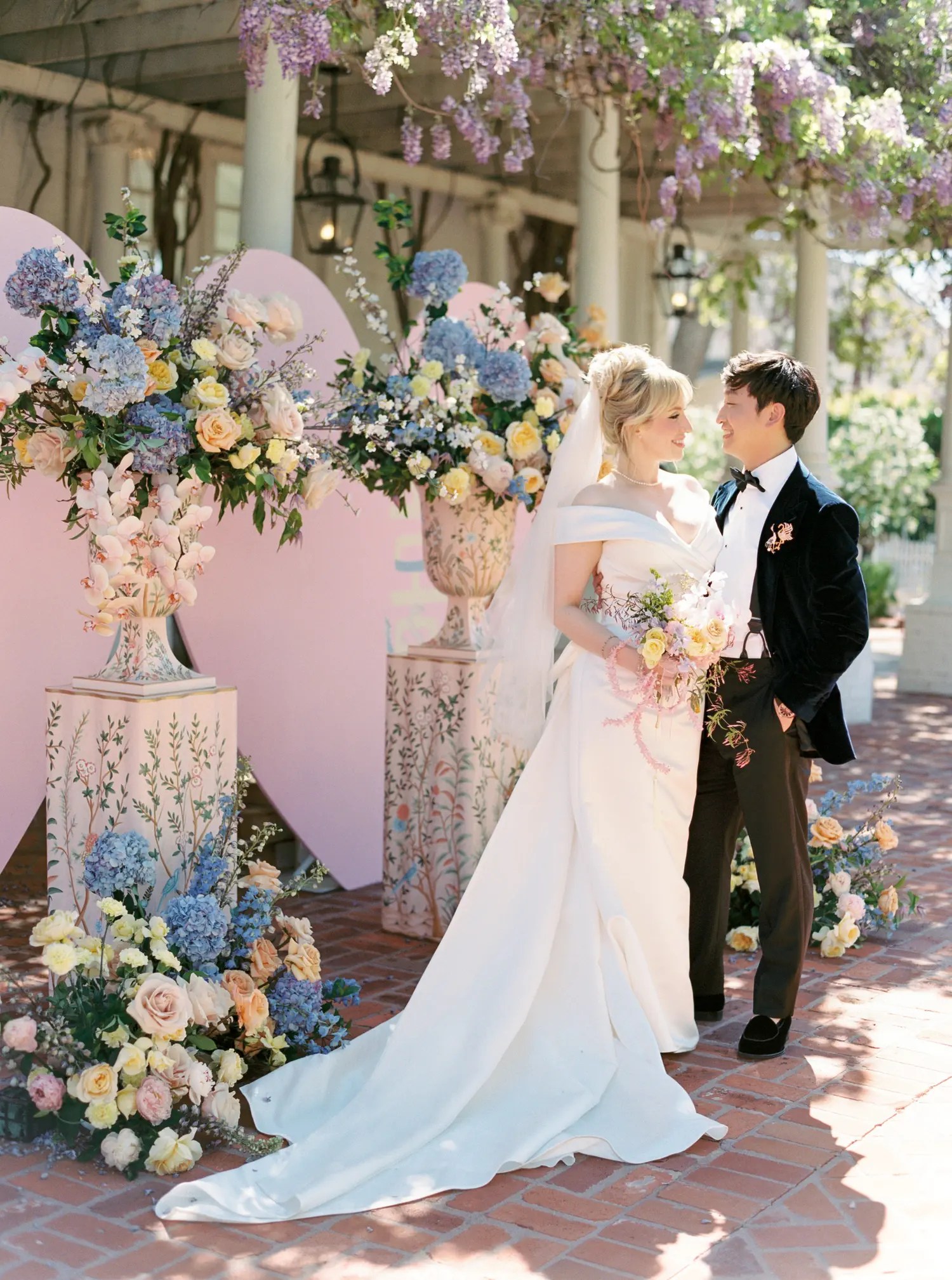 Bride holding peach and blush rose bouquet with groom beside a hand-painted pedestal urn of hydrangea and roses under wisteria